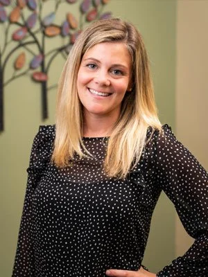 A smiling woman with long blonde hair, wearing a black polka dot dress, standing indoors against a green wall with abstract art.