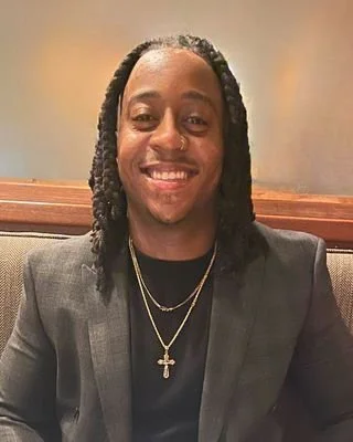 Smiling man with long braided hair wearing a dark blazer, black shirt, and layered gold necklaces with a cross pendant.