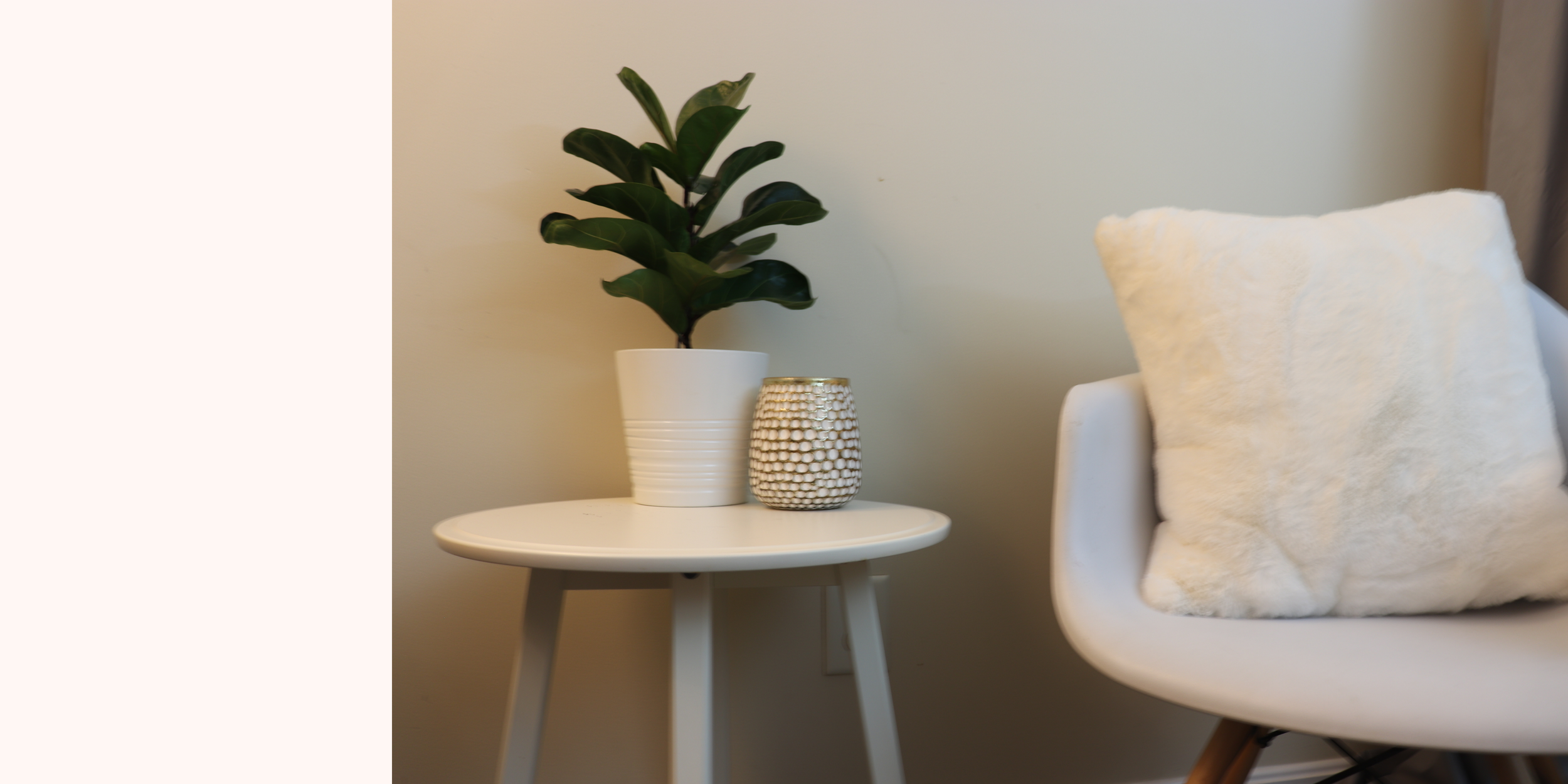 A white round side table with a potted green plant and a textured white decorative item, next to a white chair with a fluffy cream pillow in a minimalist room.