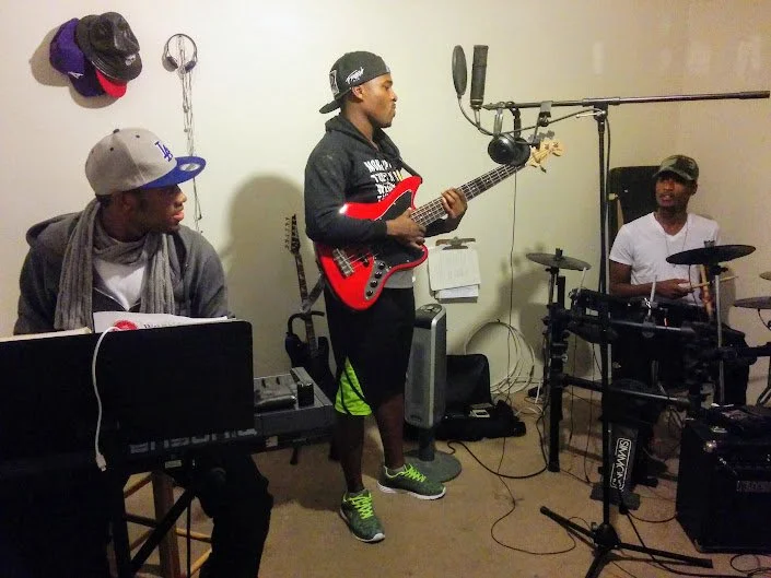 Three young men practicing music in a room with musical equipment.