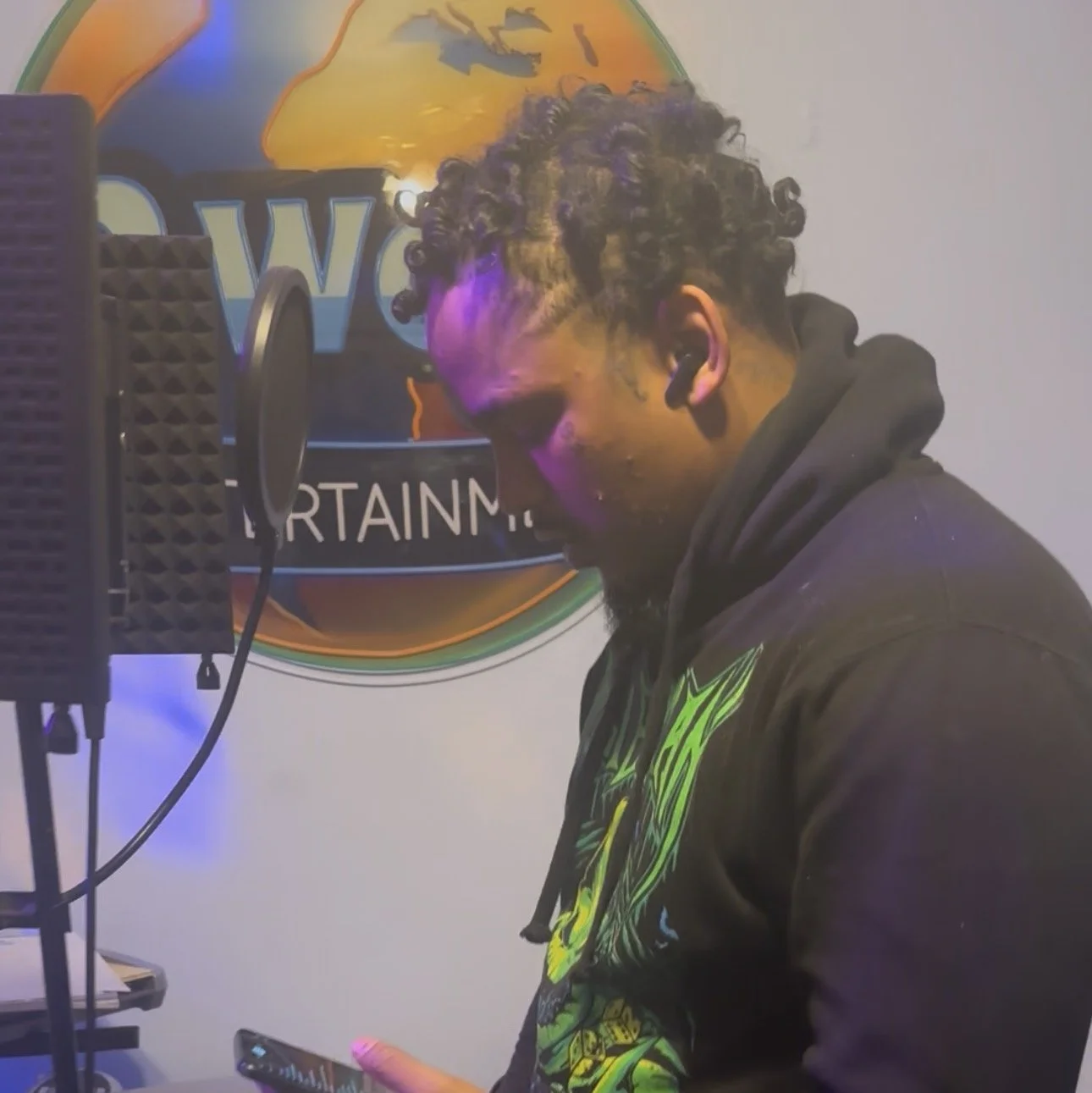 A man with curly hair and a beard, wearing earphones and a black hoodie with green and yellow graphics, stands in front of a recording microphone and shield, looking at his phone in a studio with a World of Warcraft themed sign on the wall.