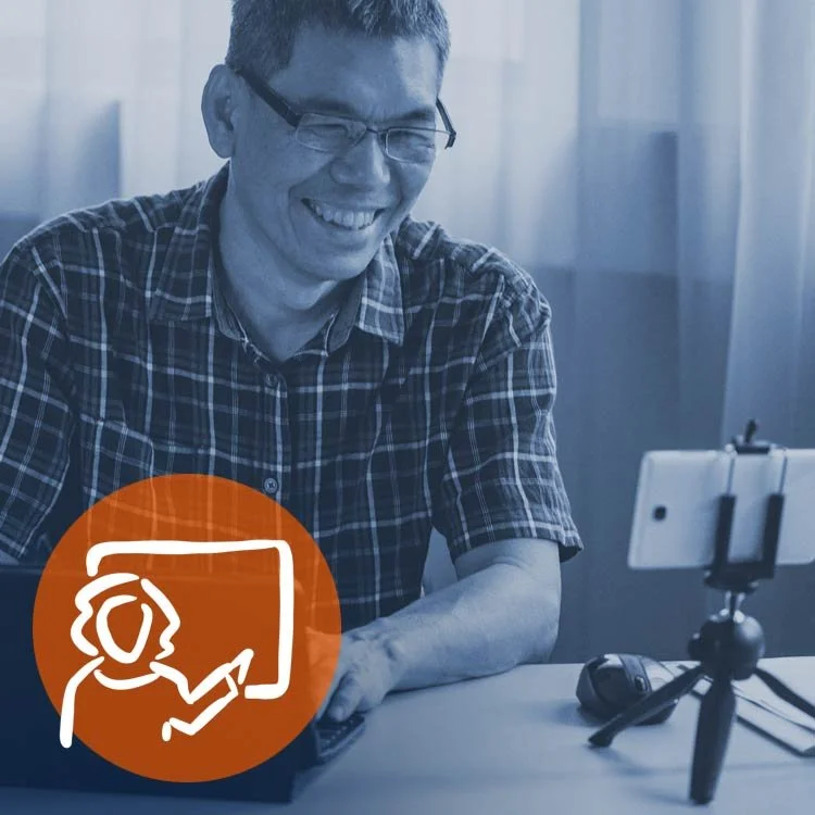 A man with glasses smiling at a laptop in a home office with a smartphone on a tripod and a computer mouse nearby.