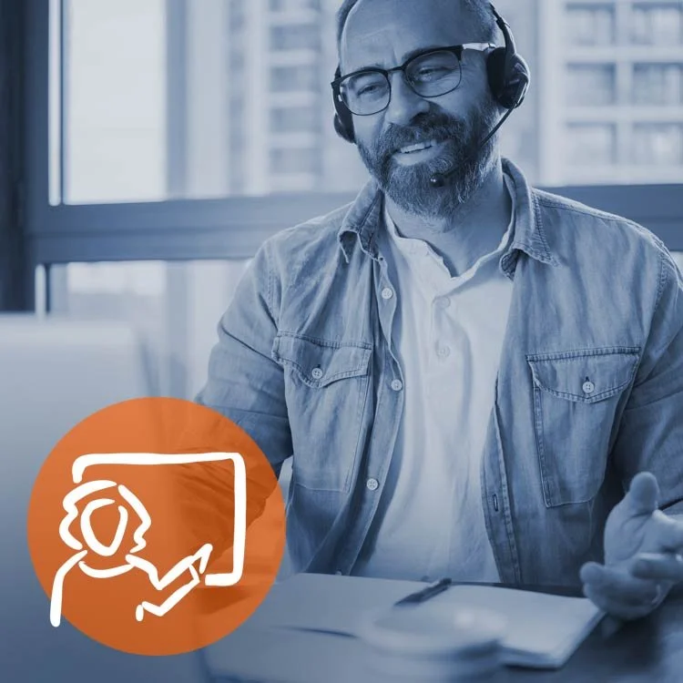 A man with glasses, a beard, and a headset sitting at a table, smiling during a video call or online meeting.