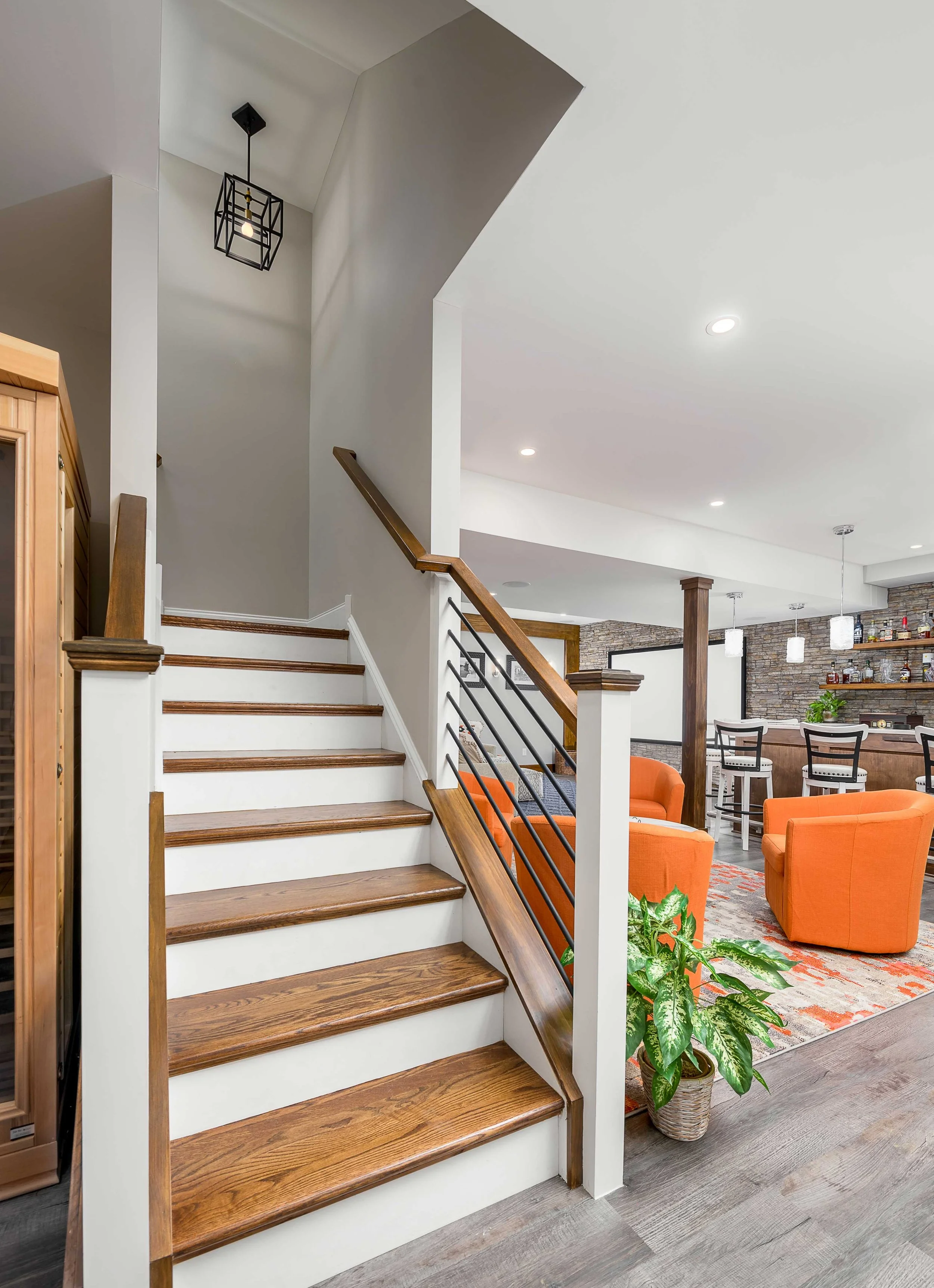 Interior view of a modern home featuring a staircase with wooden steps and a metal and wood railing. In the background, there is a living area with orange armchairs, a rug, a potted plant, and a stone accent wall with shelves and decor.