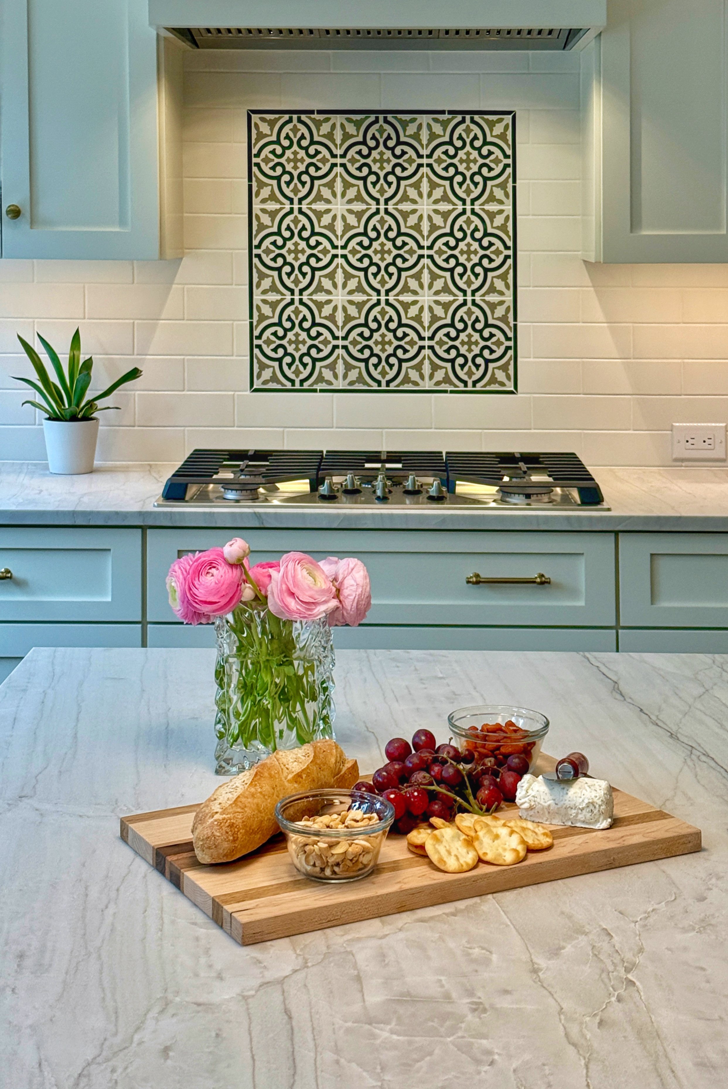 A kitchen scene with a white marble countertop, a vase with pink flowers, a wooden cutting board with bread, grapes, cheese, and snacks. In the background, a decorative tile backsplash, a potted plant, and a stove.