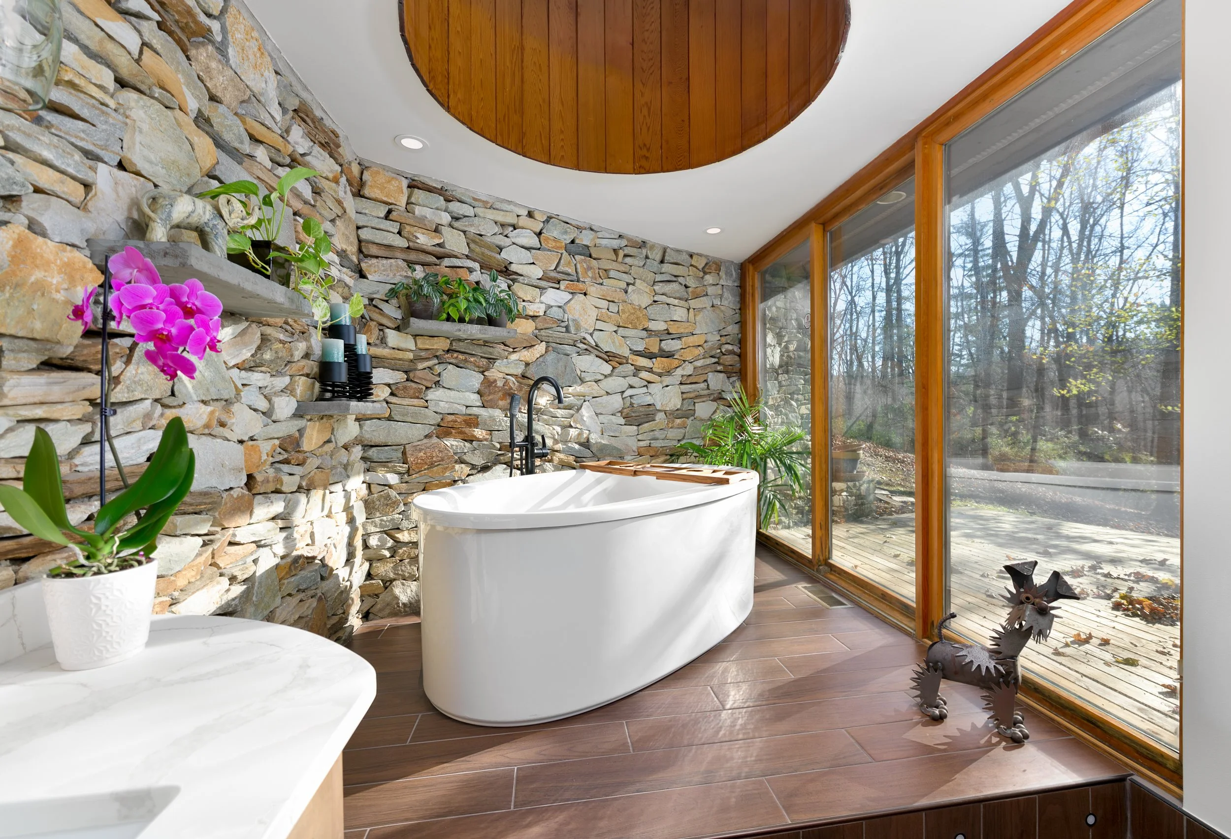 Luxurious bathroom with stone walls, a freestanding bathtub, large windows, and plants.
