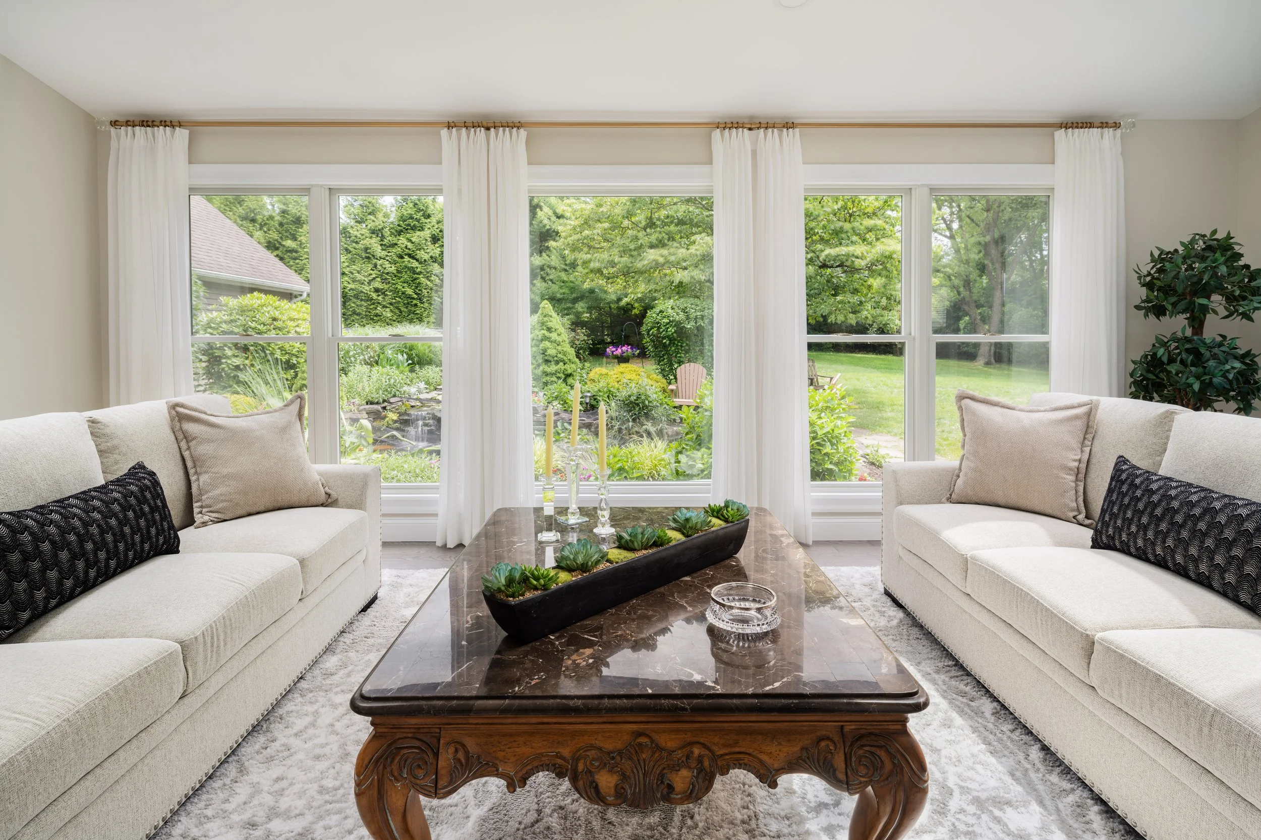 Bright living room with white sofas, marble coffee table, large windows with white curtains, and garden view outside.