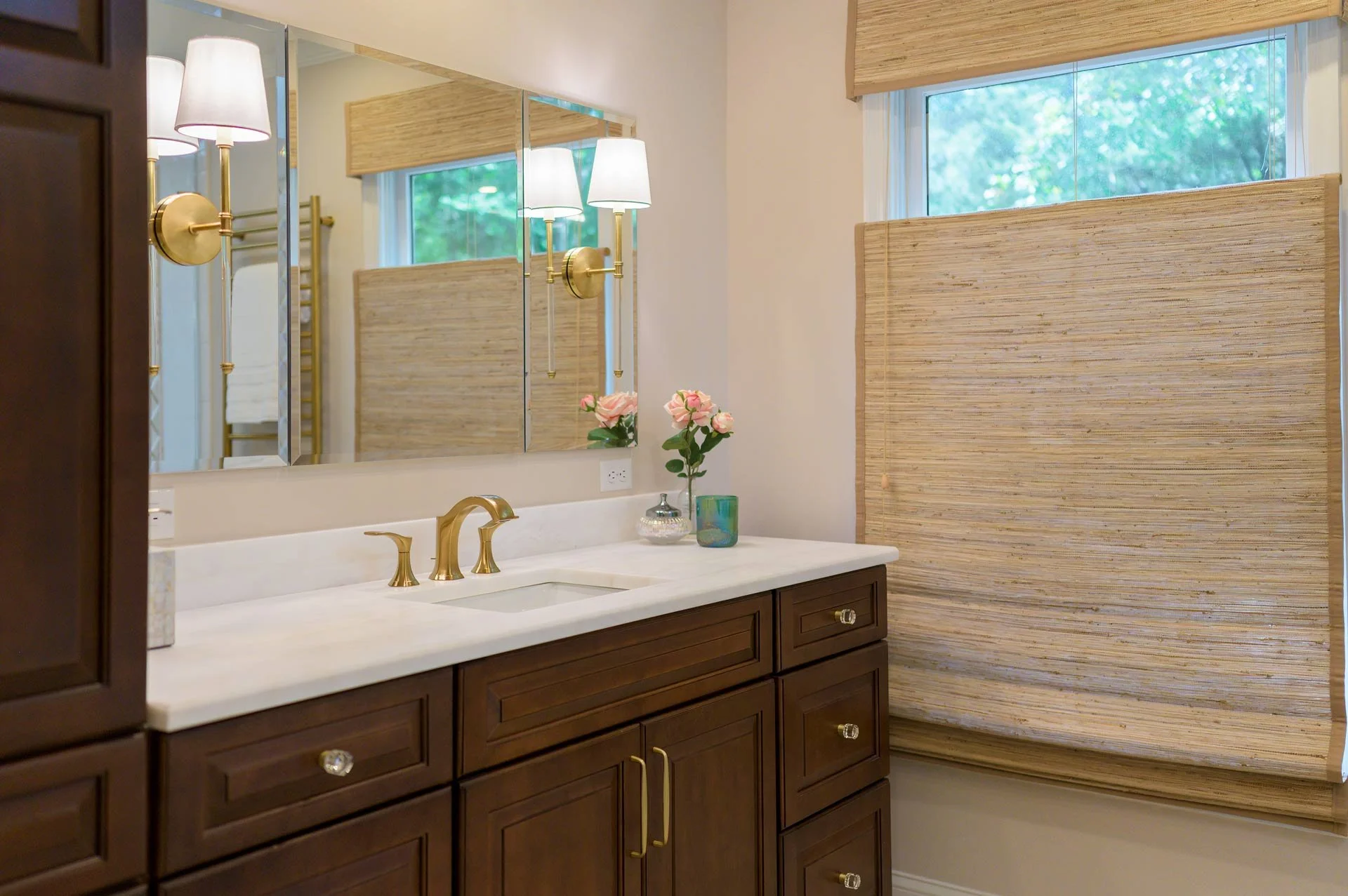 Modern bathroom with wooden cabinets, white countertop, gold faucet, mirror, wall sconce, floral decor, and window with bamboo shades.