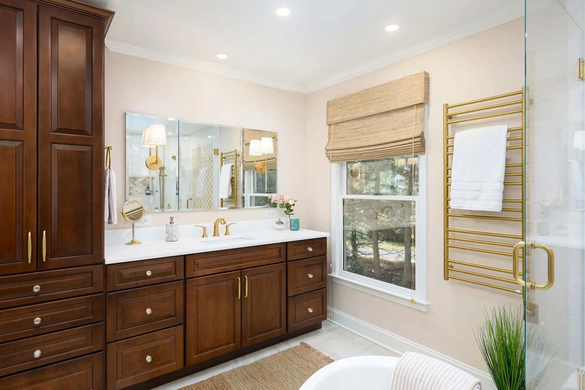 Modern bathroom with wooden cabinets, marble countertop, gold fixtures, mirror, towel rack with white towel, window with bamboo shade, and decorative plants.