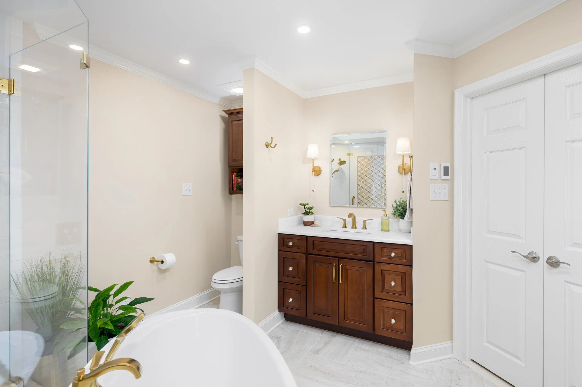 Modern bathroom interior with a wooden vanity, white countertop, and gold fixtures. There is a mirror above the sink with wall-mounted lights on either side. A toilet is visible next to a glass shower enclosure. Potted plants are placed for decoratio