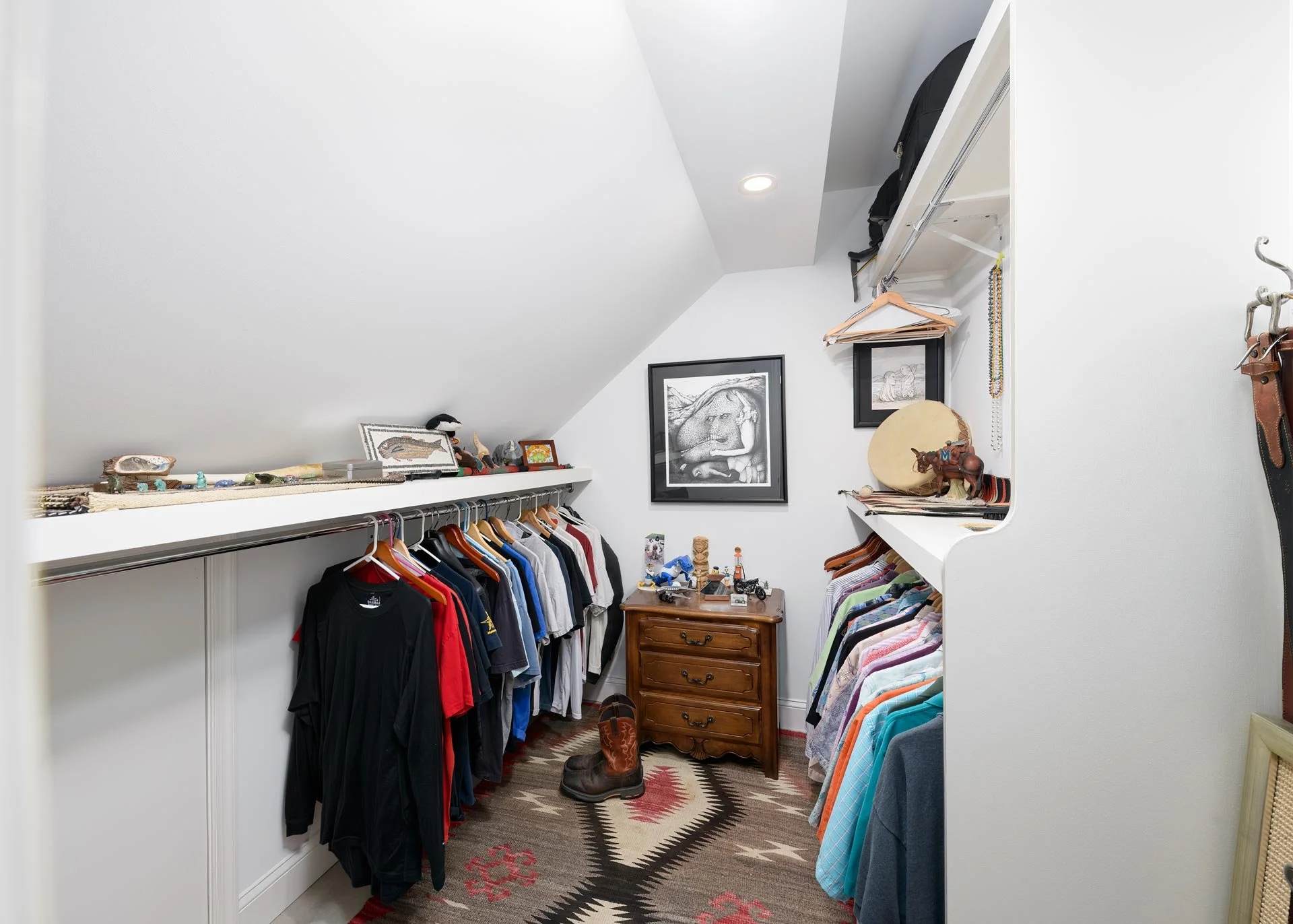 Walk-in closet with clothes hanging on racks, a small wooden dresser, decorative items on shelves, framed artwork on walls, and a patterned carpet on the floor.