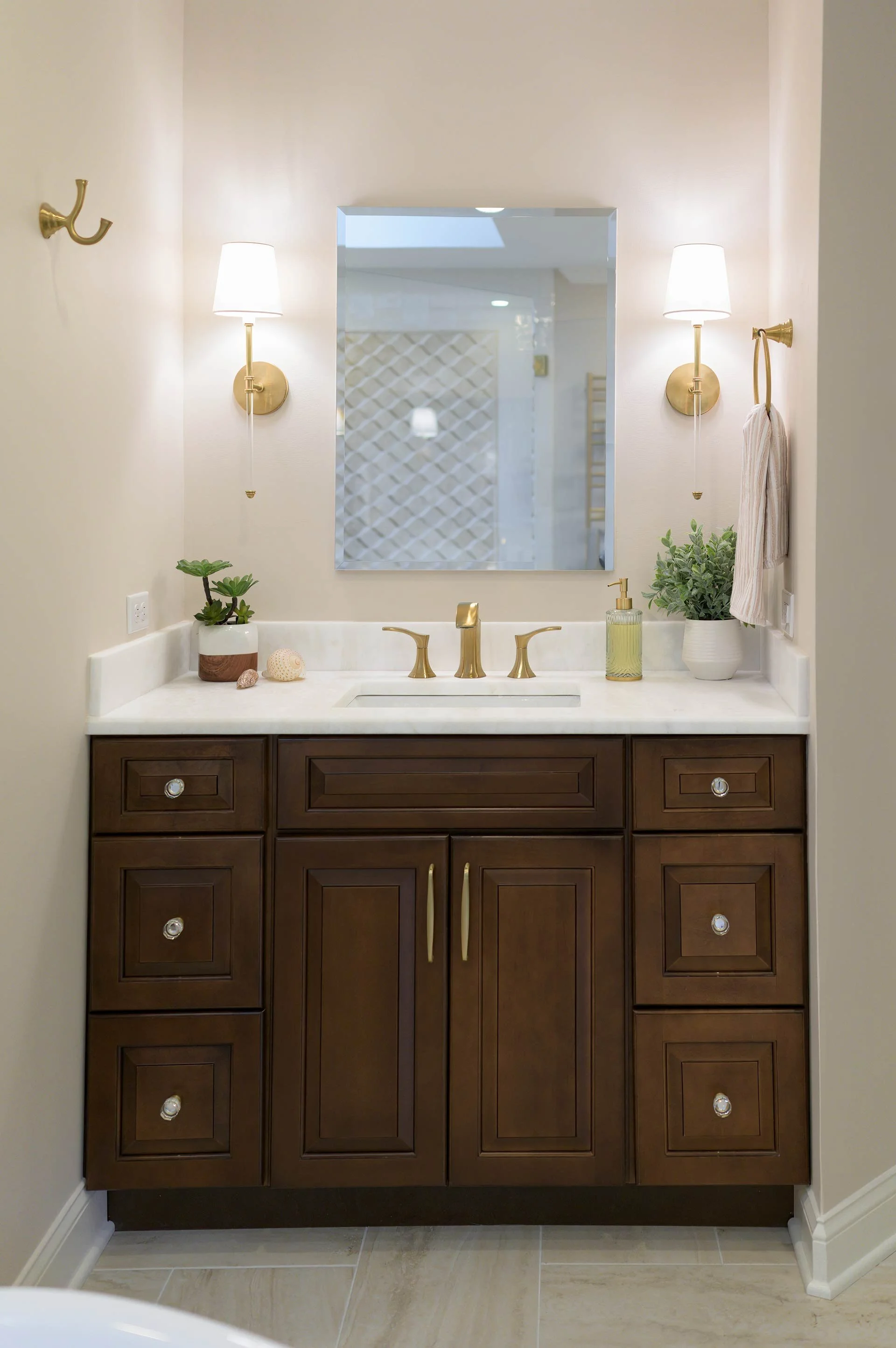 A modern bathroom vanity with a marble countertop, brown cabinets, gold faucet, and two wall-mounted lights. A mirror is centered above the sink. The countertop holds a plant, seashells, and a soap dispenser.