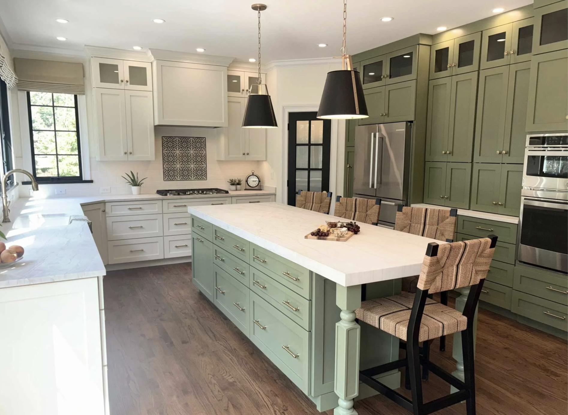 Modern kitchen with white and green cabinets, a large white island, black pendant lights, and stainless steel appliances. There's a window with a view outside and a dining area with upholstered chairs.