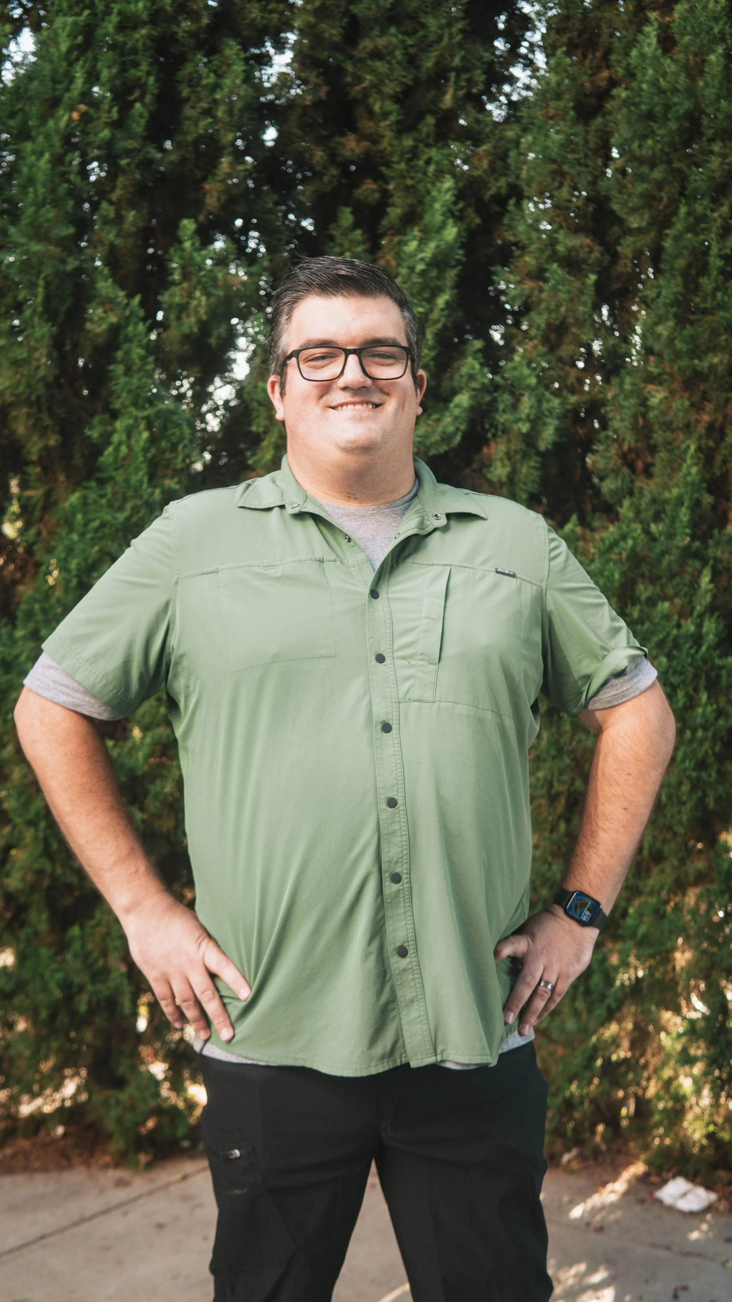 A man with glasses, dark hair, and light skin is standing outside in front of green bushes, smiling and with hands on hips. He is wearing a light green short-sleeve shirt over a gray t-shirt, black pants, and a smartwatch on his left wrist.