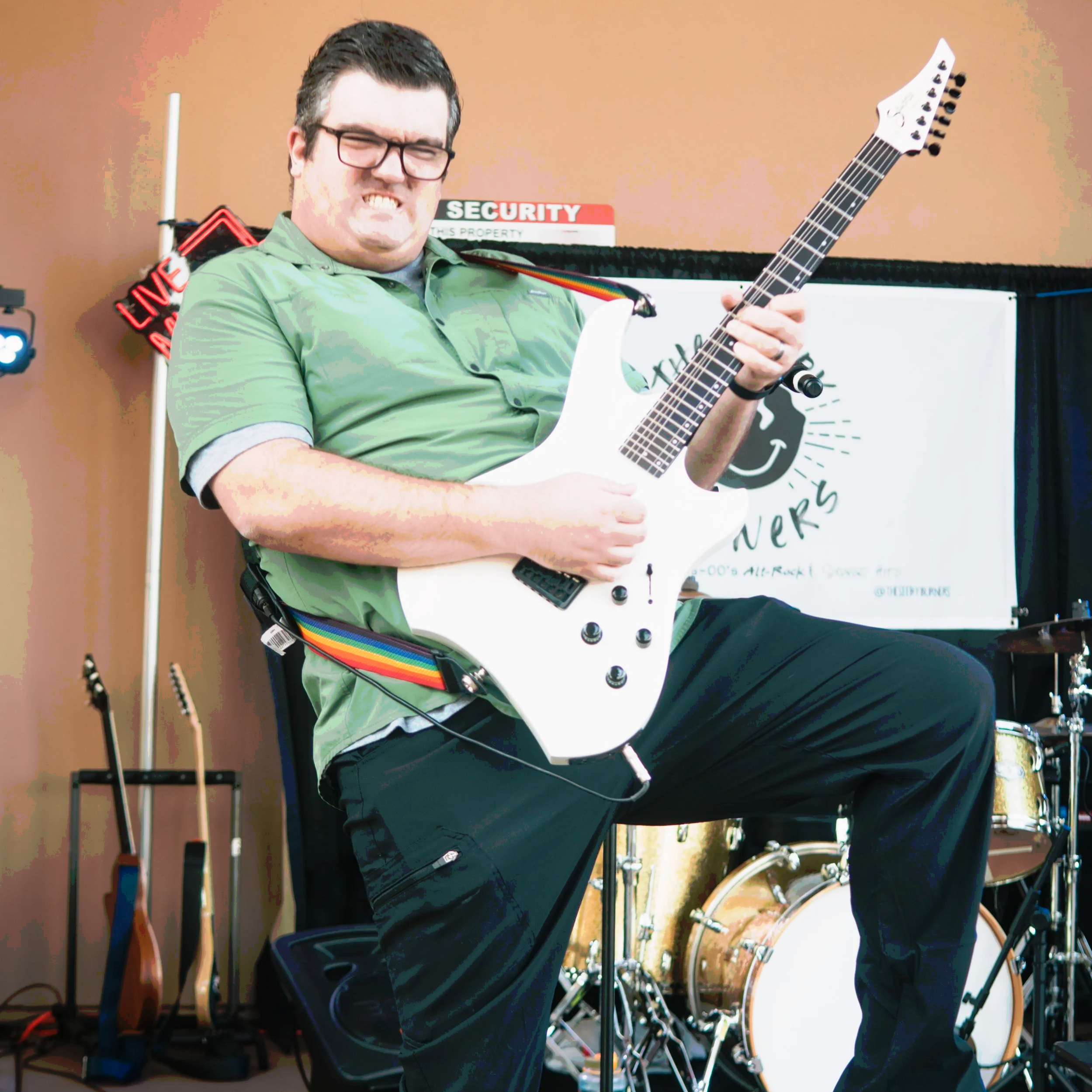 Man playing electric guitar on stage with musical instruments and a banner in the background.