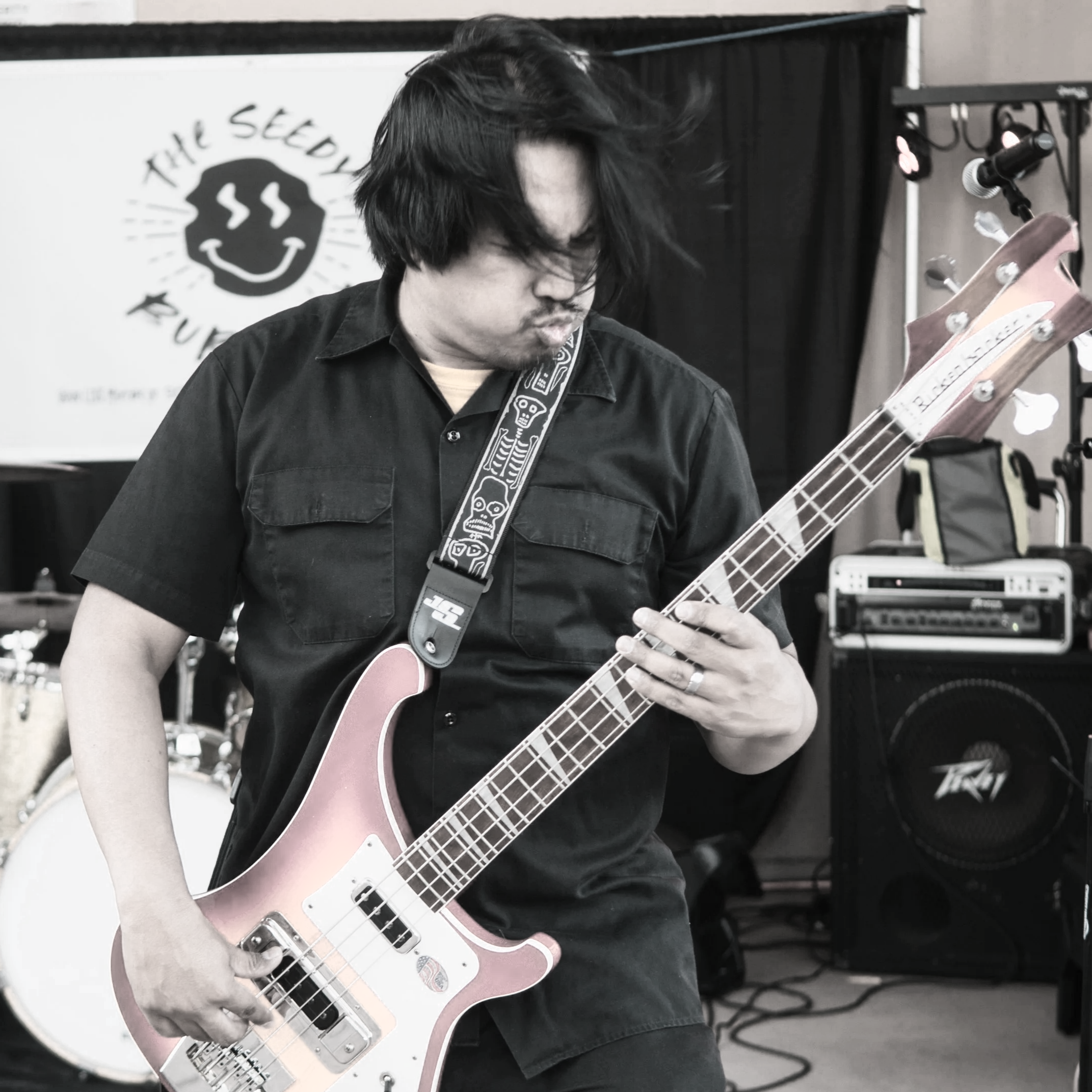 A man with black hair playing an electric bass guitar in a music venue, with performance equipment in the background.