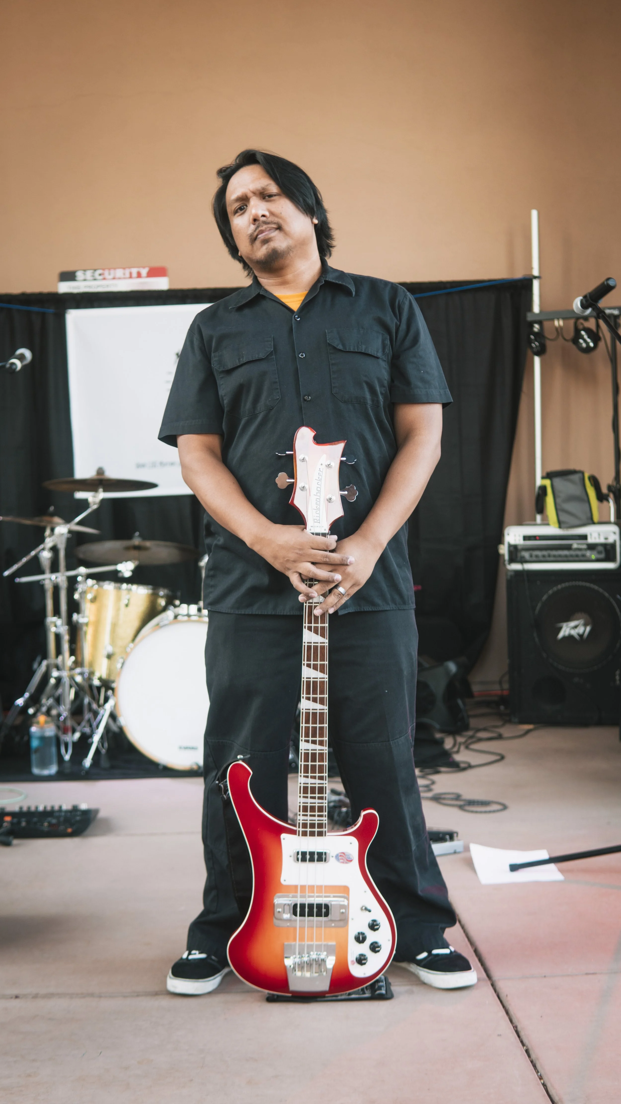 A man in black clothing holding a red and white electric bass guitar stands on a stage with musical instruments and equipment in the background