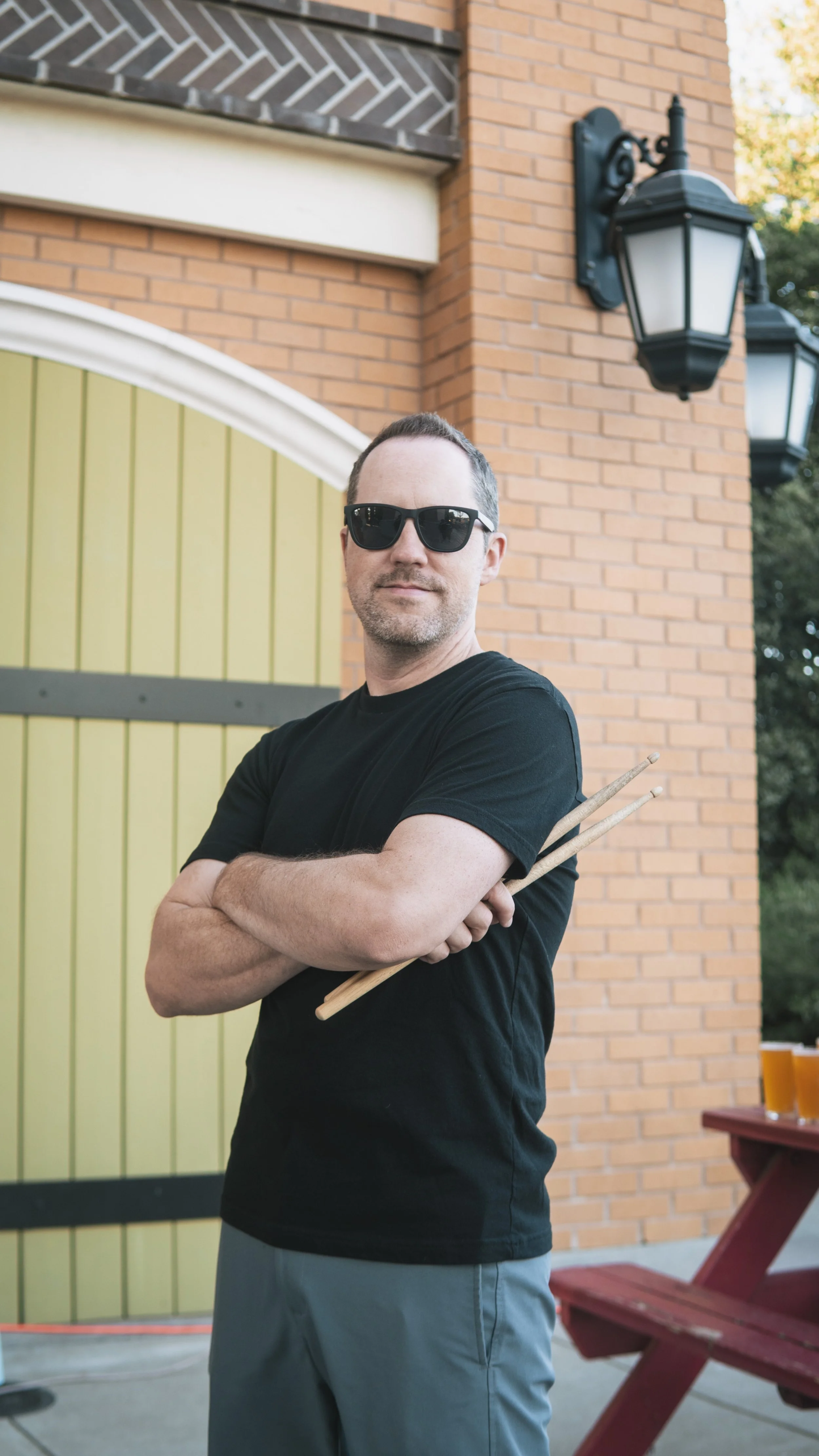 A man wearing black sunglasses and a black t-shirt, standing outdoors with arms crossed, holding wooden chopsticks against a backdrop of a brick building with hanging lanterns and a yellow-green gate.