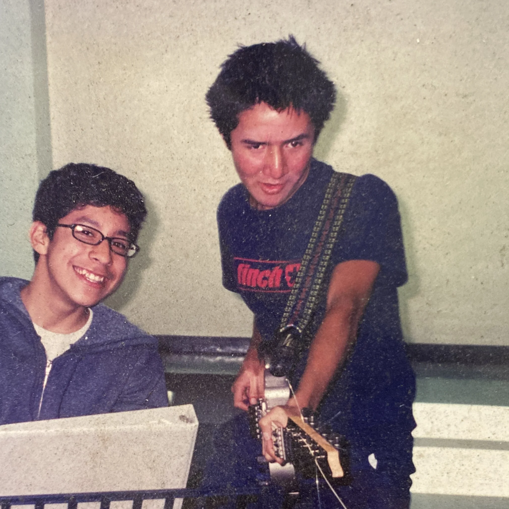 Two young men in a casual setting, one sitting at a table with a sheet of paper, smiling with glasses, and the other standing, holding an electric guitar, wearing a black T-shirt with red text.