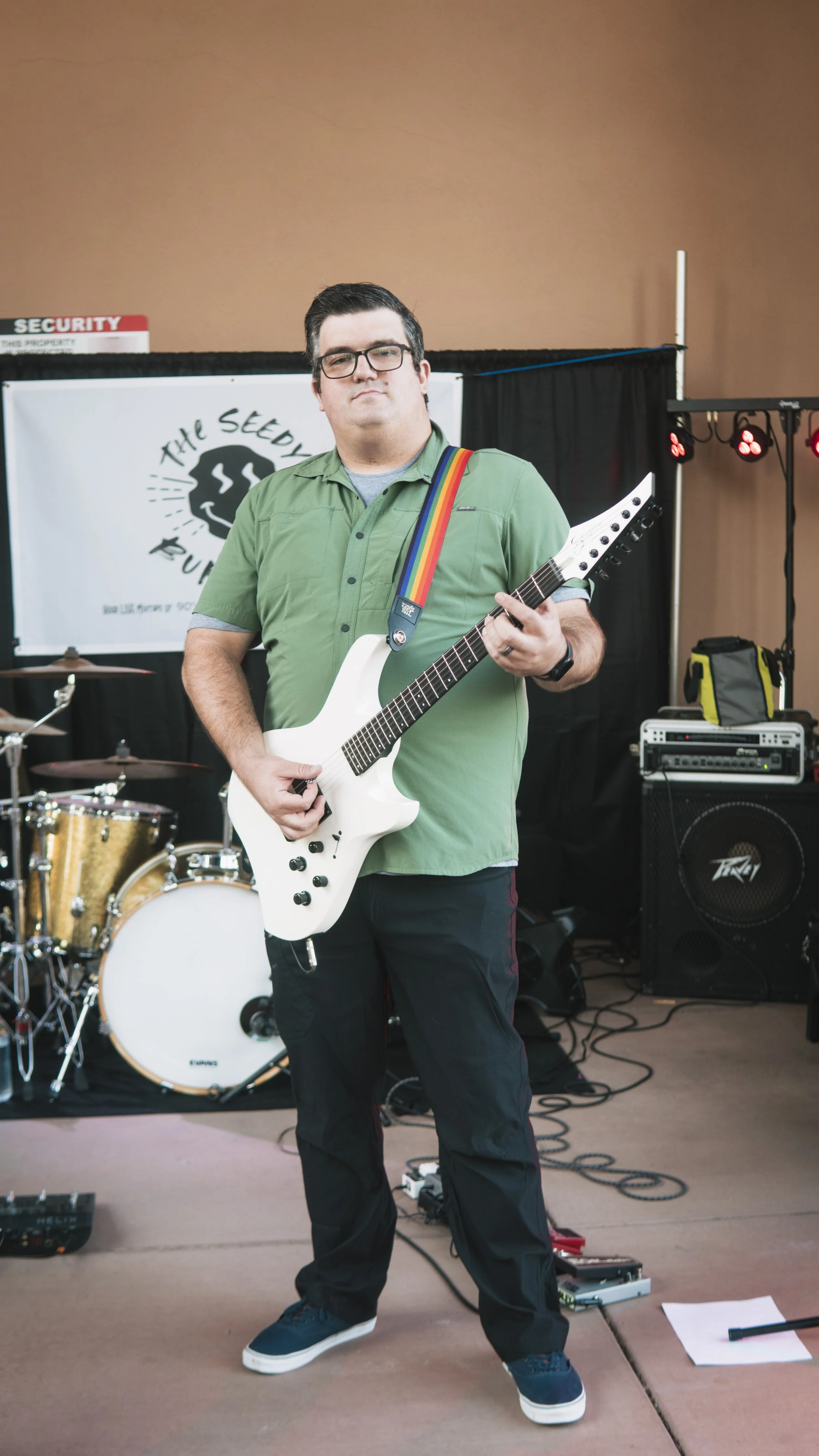 A man in a green shirt and black pants holding a white electric guitar on stage. There is a drum set and sound equipment behind him, with a black curtain and a banner that says 'The Seedle...' in the background.