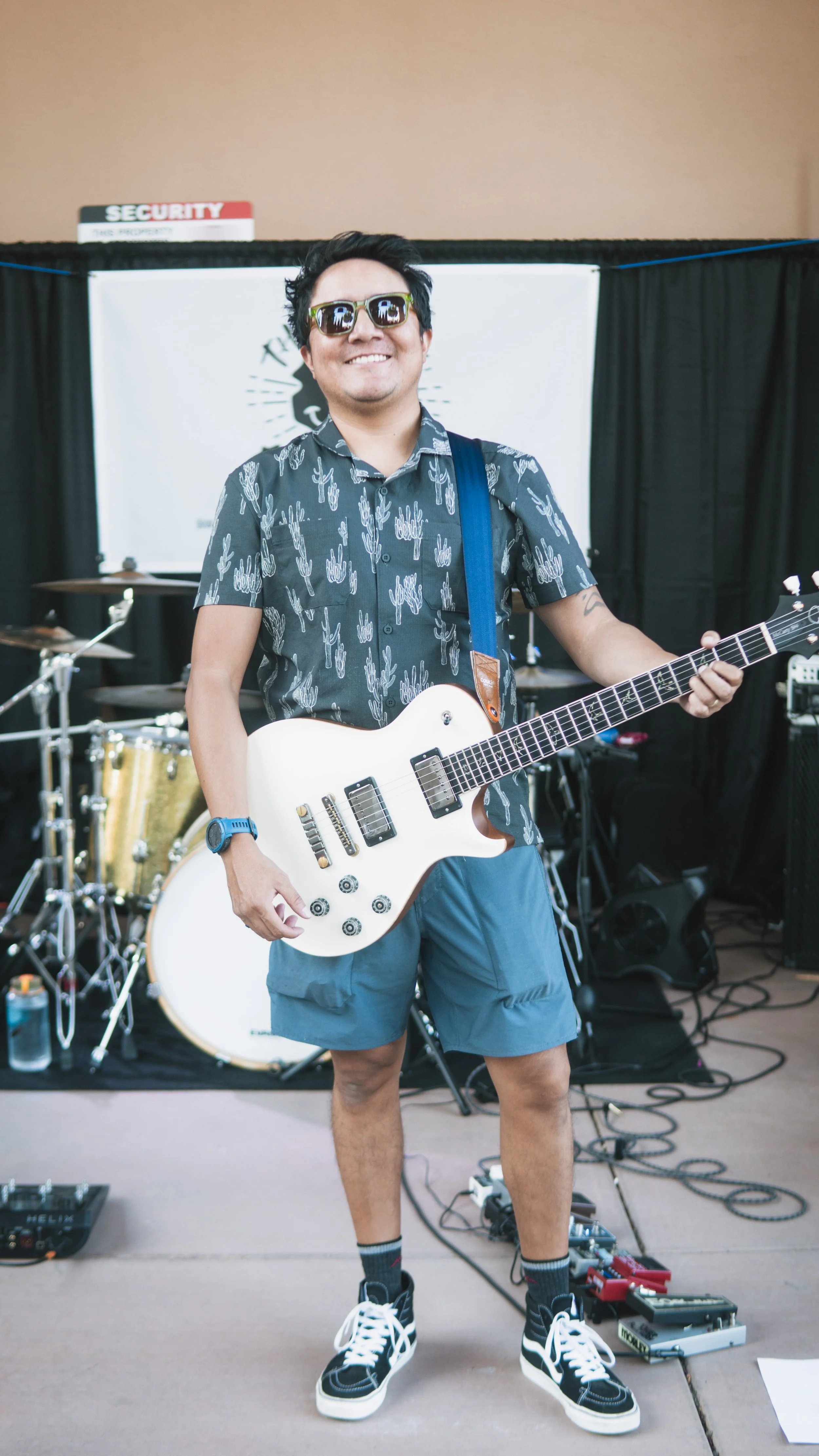 Smiling man wearing sunglasses, a button-up shirt with cactus and succulent patterns, blue shorts, and sneakers, holding a white electric guitar, standing on a stage with musical equipment behind him.