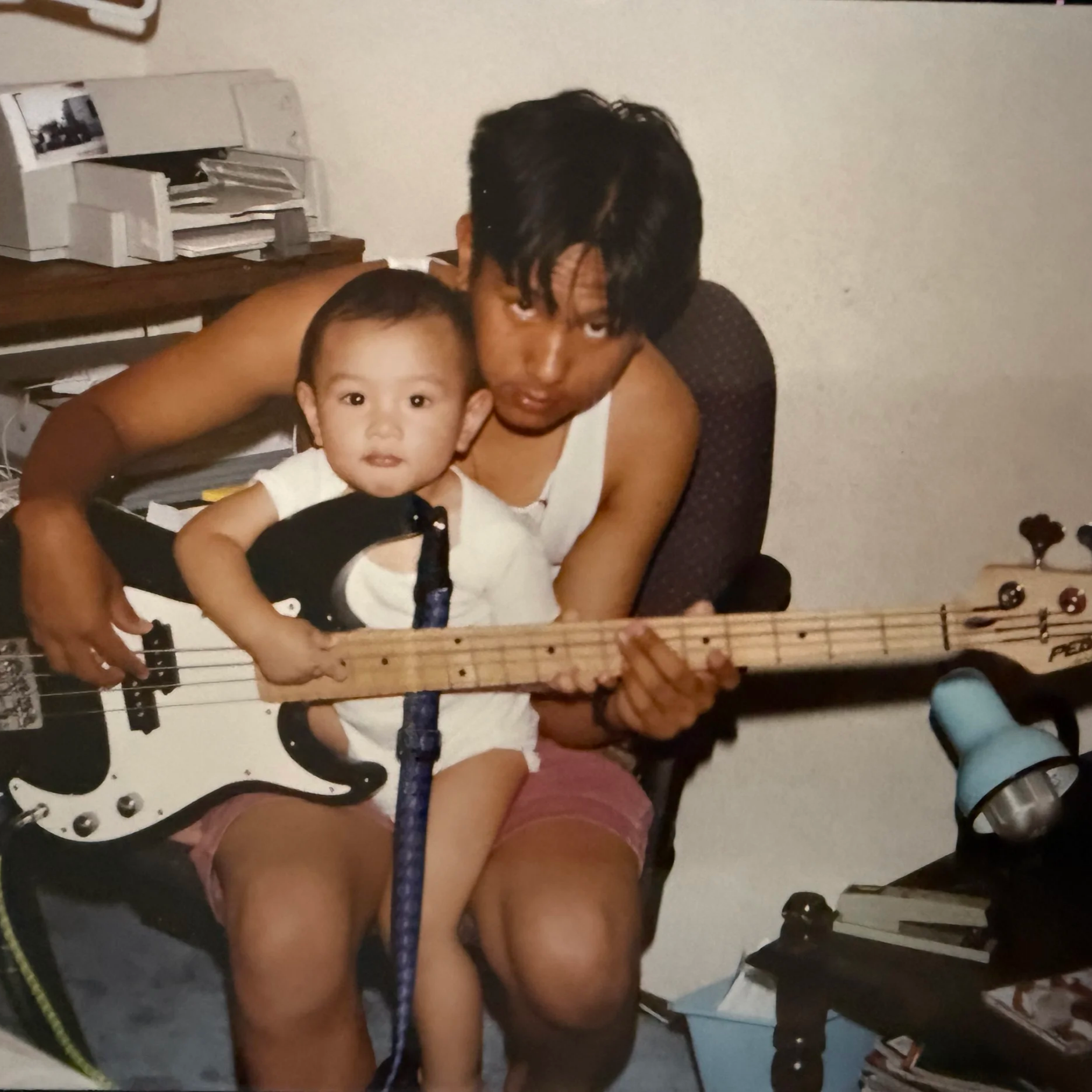 A man and young boy sitting together in a room, the man holding a bass guitar and the boy sitting on his lap, looking at the camera.