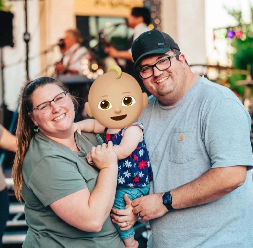 Family posing and smiling with a woman, man, and a toddler in an outdoor setting, blurred background with people and lights.