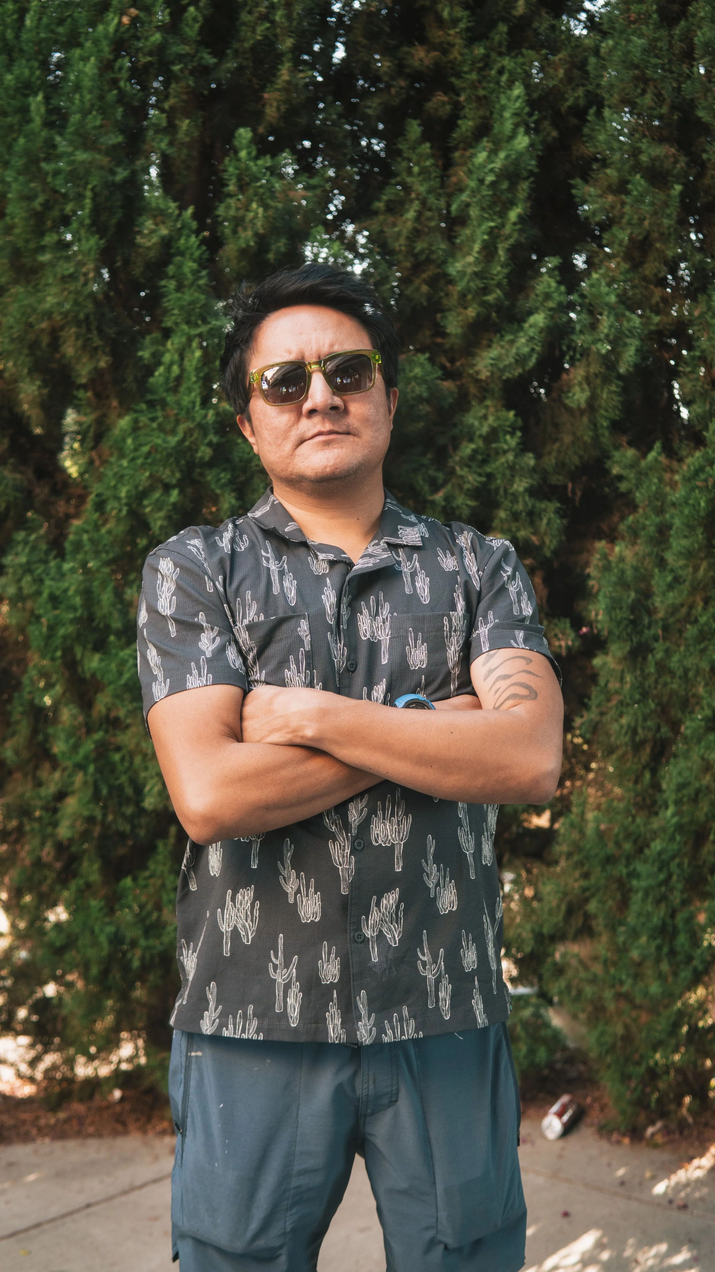 A man with crossed arms wearing sunglasses, a black short-sleeved shirt with cactus patterns, and dark shorts, standing outdoors in front of green bushes.