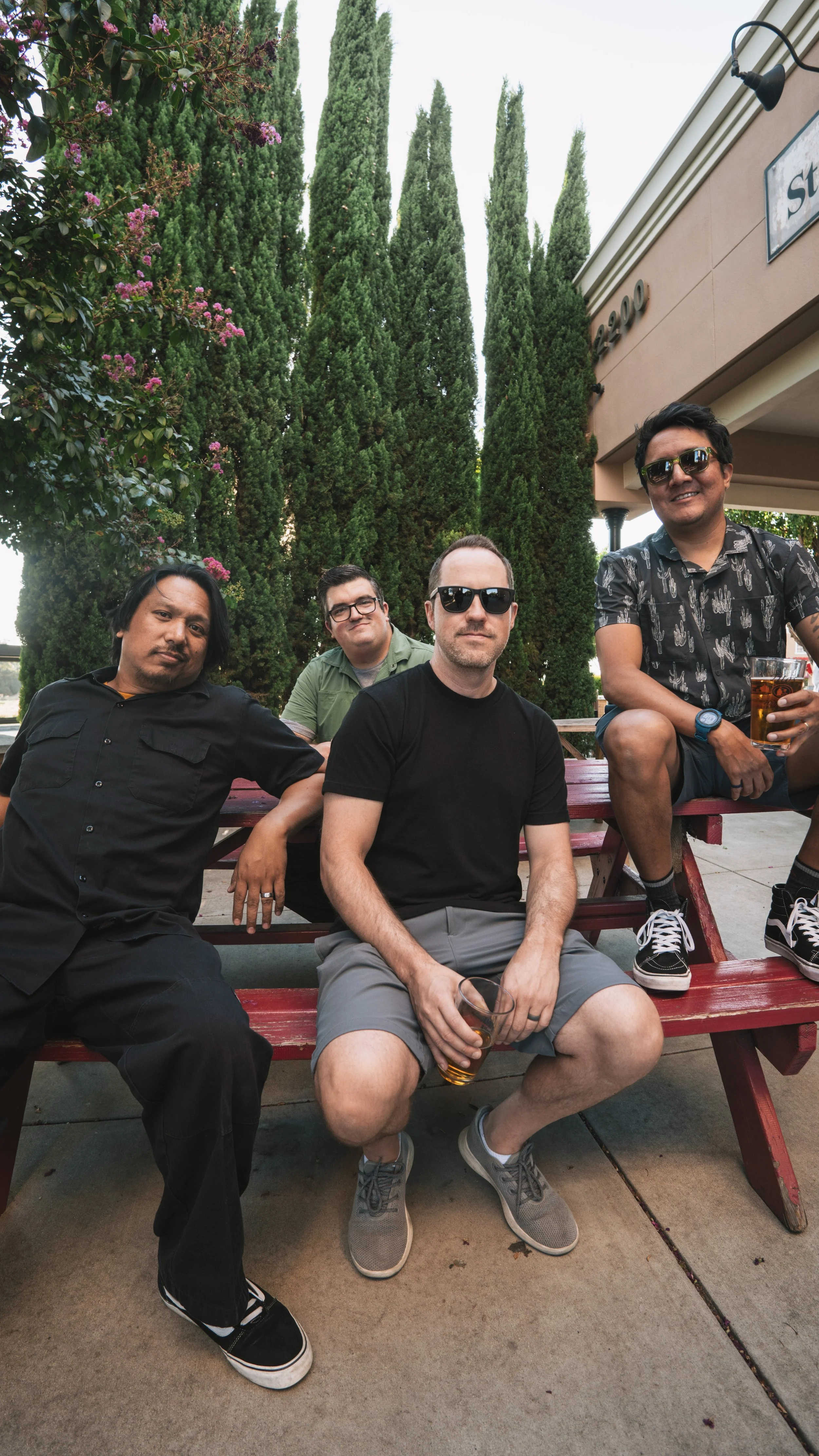 Four men sitting and standing around a red picnic table outside, with tall green trees and a building in the background, holding drinks and smiling.