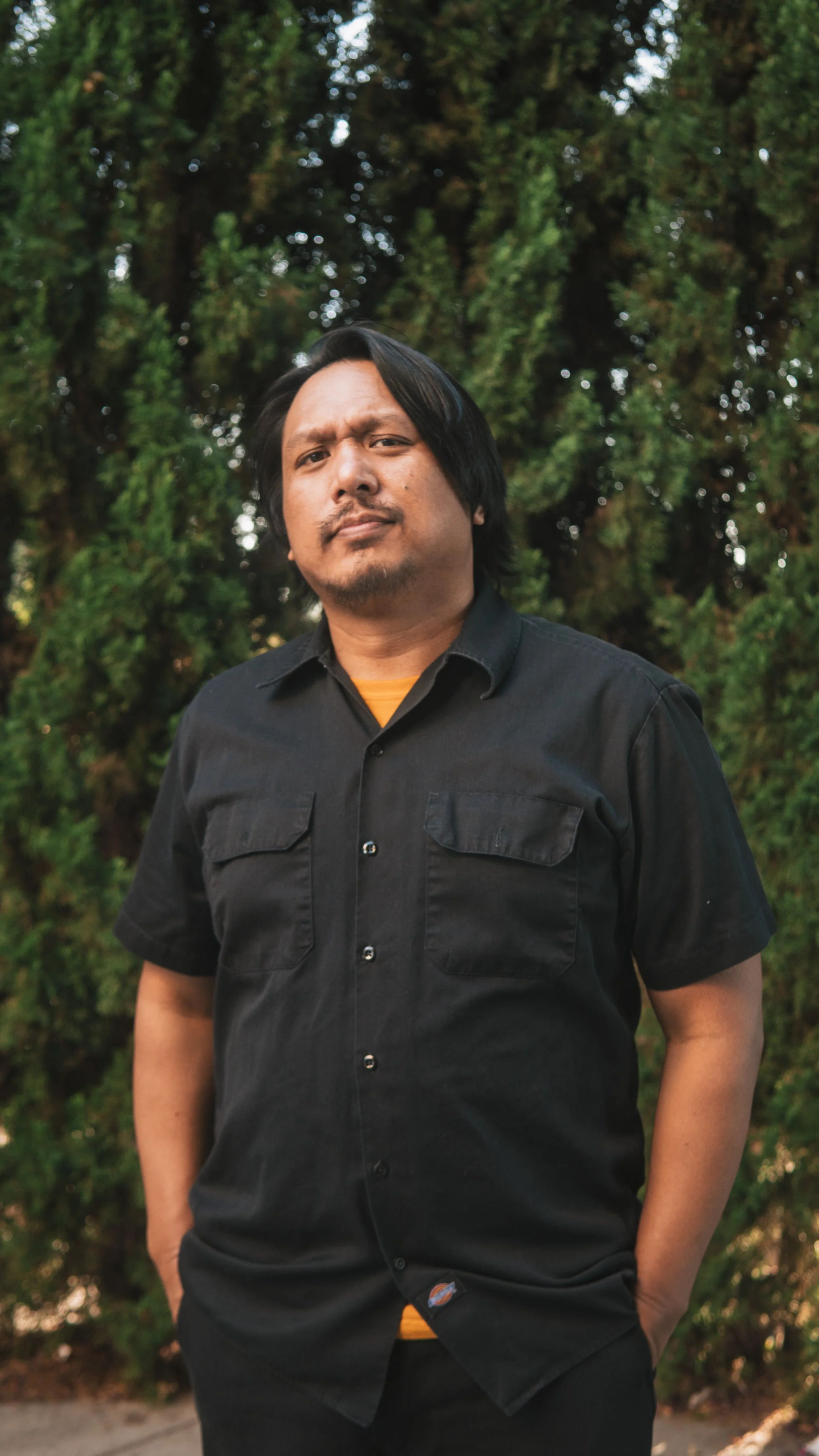 A man standing outdoors in front of green trees, wearing a black short-sleeved button-up shirt and looking at the camera with a serious expression.