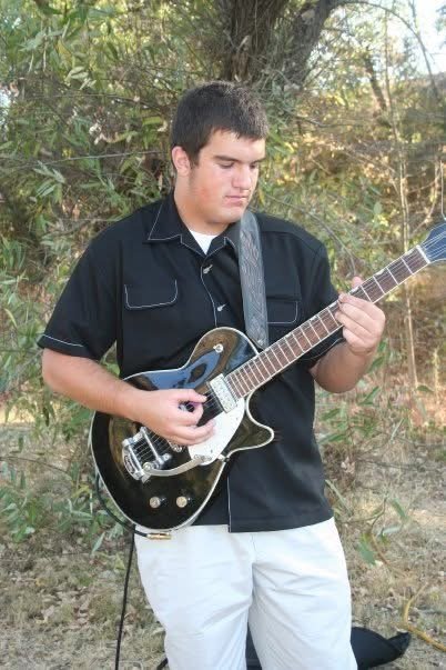 Young man playing an electric guitar outdoors near trees and bushes.