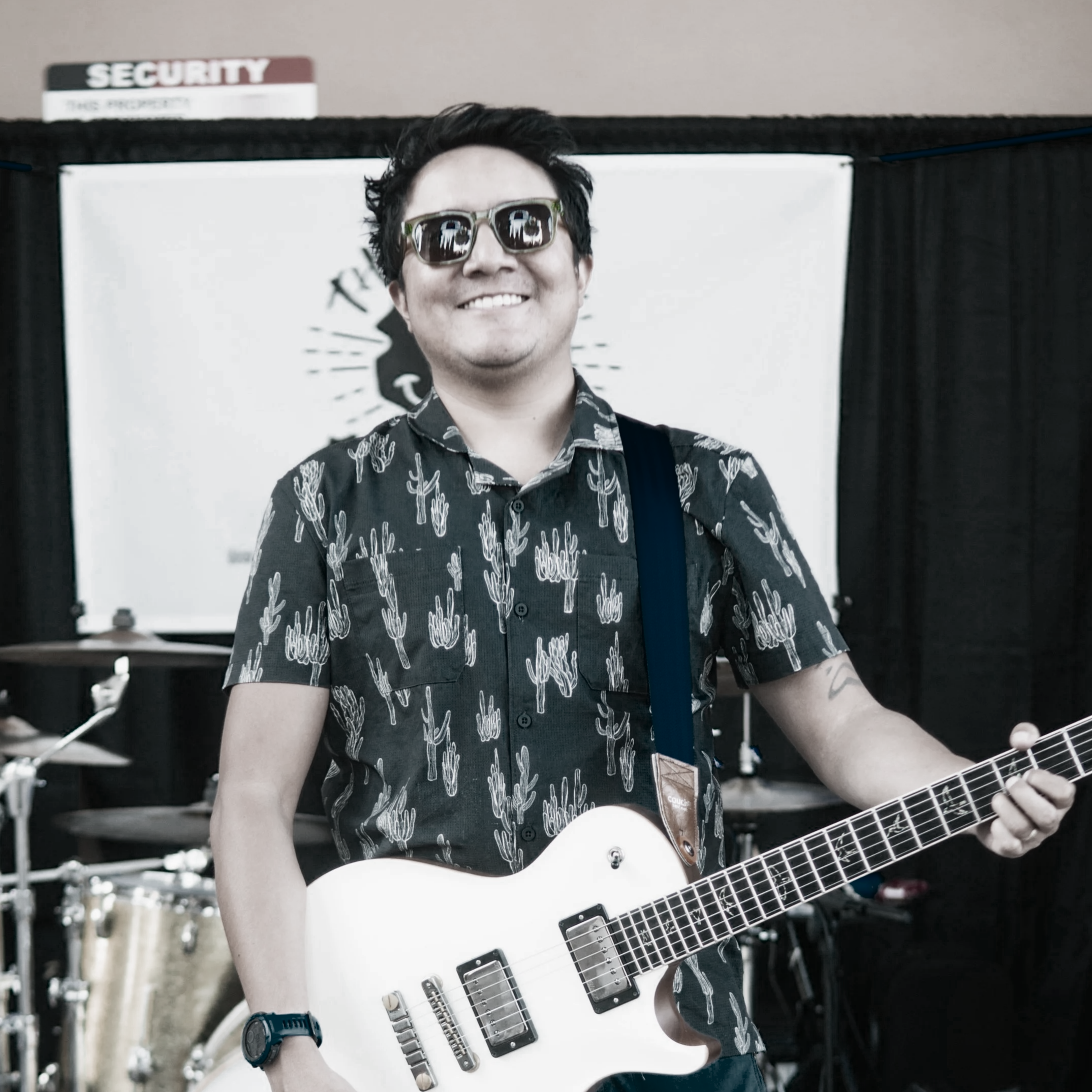 A man smiling and wearing sunglasses, holding a white electric guitar, standing in front of a black curtain with the silhouette of a lion.