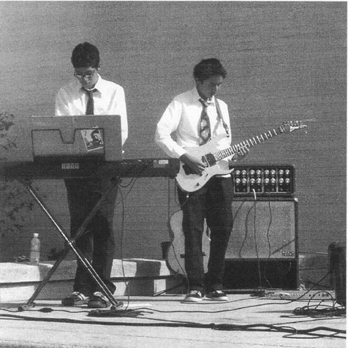 Two young men performing music on stage, one playing keyboard and the other playing electric guitar, with musical equipment surrounding them.