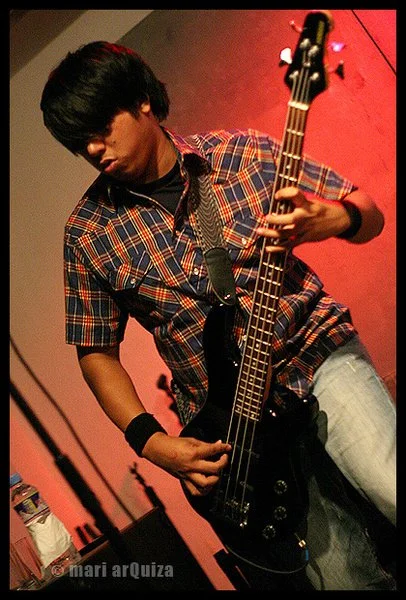 A young man with dark hair wearing a plaid shirt and jeans playing an electric bass guitar on stage.