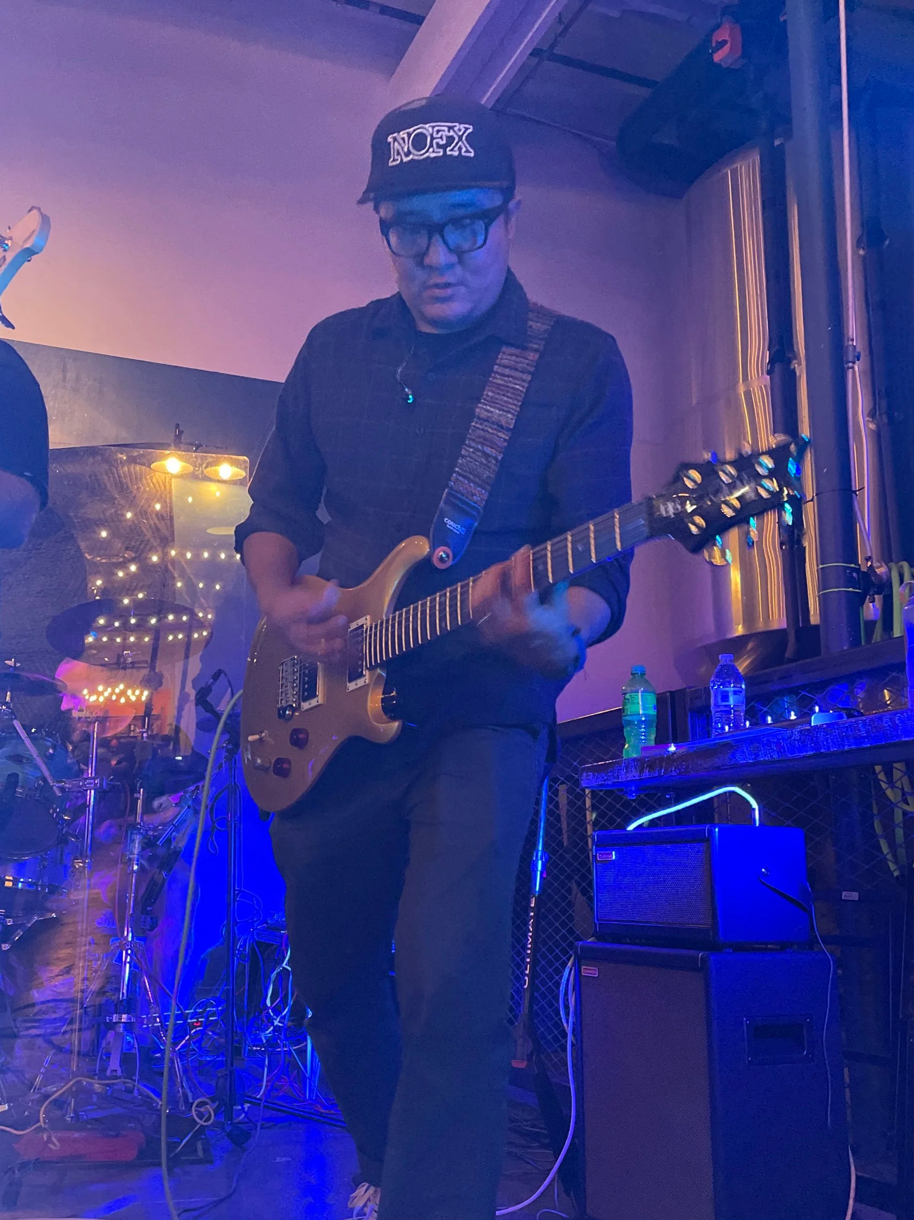 A man wearing glasses, a black cap with the word 'NOFX', and a dark shirt playing an electric guitar during a live music performance in a dimly lit venue with blue lighting.