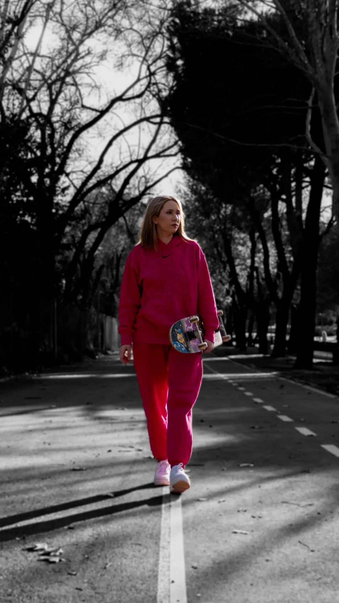 Mujer caminando por un sendero arbolado con un skateboard en la mano, vestida con un conjunto deportivo rosa intenso. La imagen destaca por su efecto artístico de color selectivo, transmitiendo estilo urbano, libertad y actitud casual.