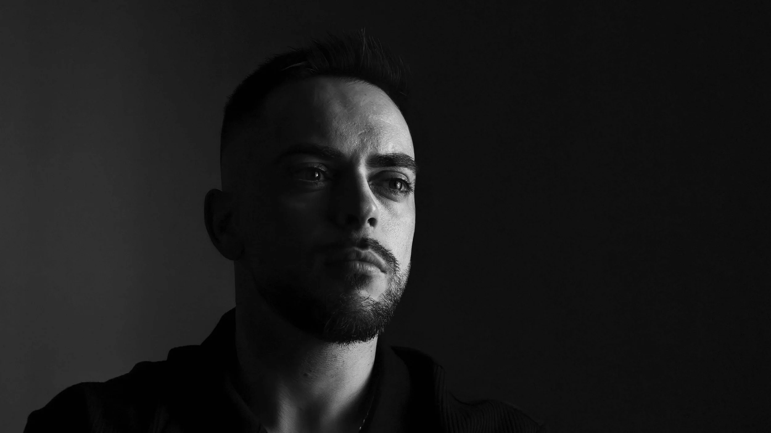 Retrato masculino en estilo low-key, capturado con iluminación lateral que resalta la textura de la piel y las facciones del rostro. La composición en blanco y negro crea un ambiente dramático y elegante, transmitiendo introspección y carácter. Fotog