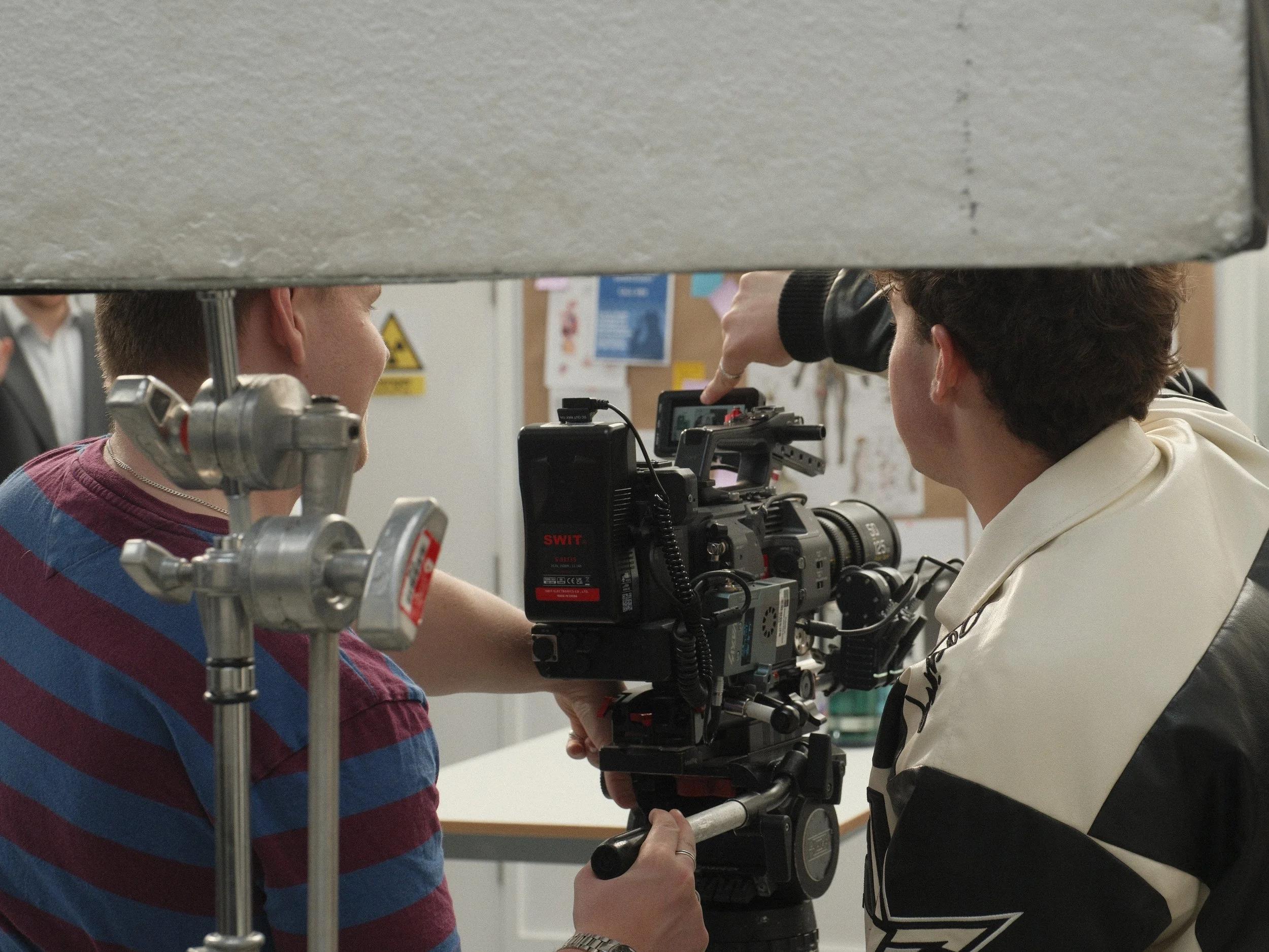 Two people operating a professional film camera behind a gray barrier with a workshop or office environment in the background.
