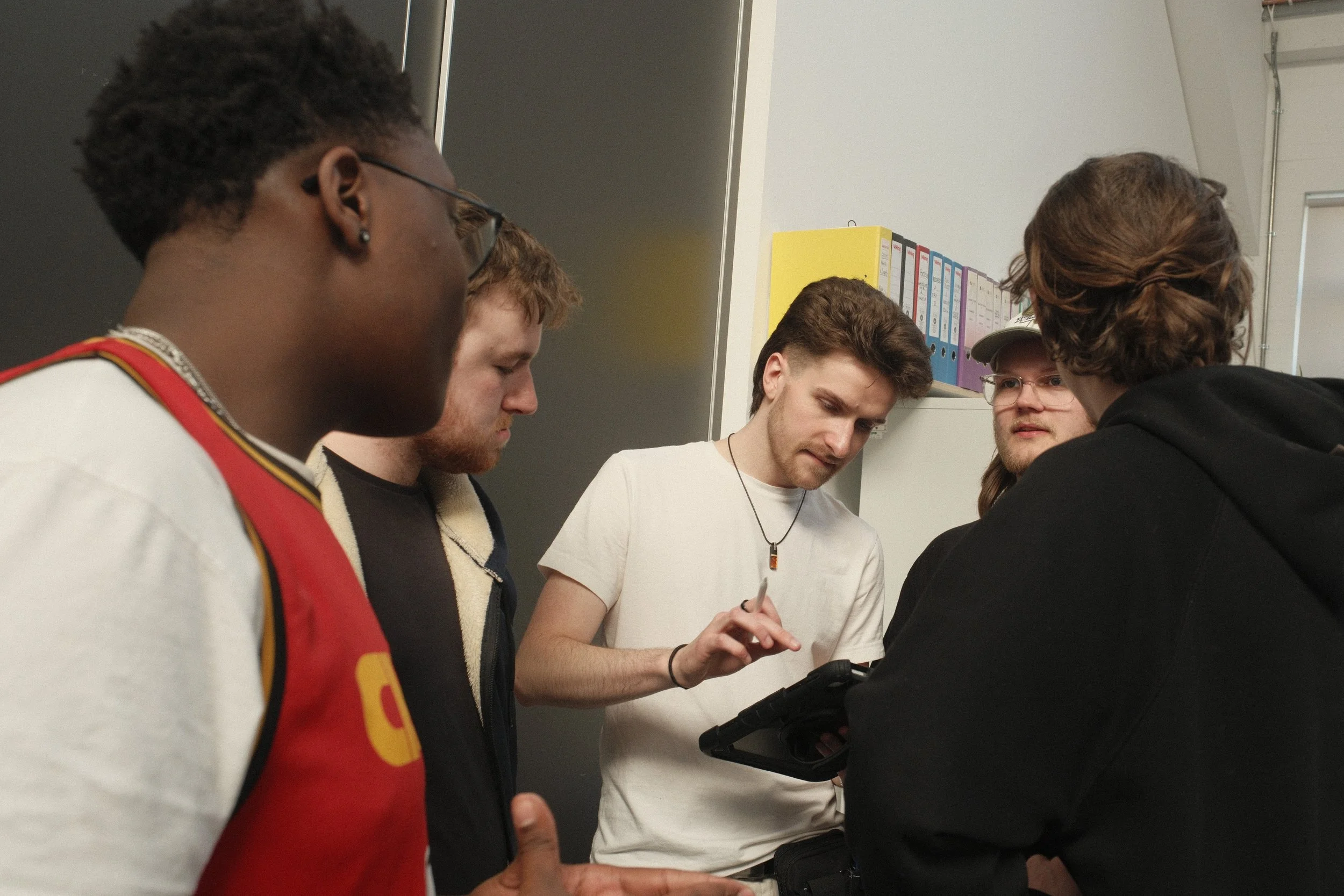 Five young men gathered, focusing on a tablet device held by one of them, in an indoor setting with shelves of colorful binders in the background.