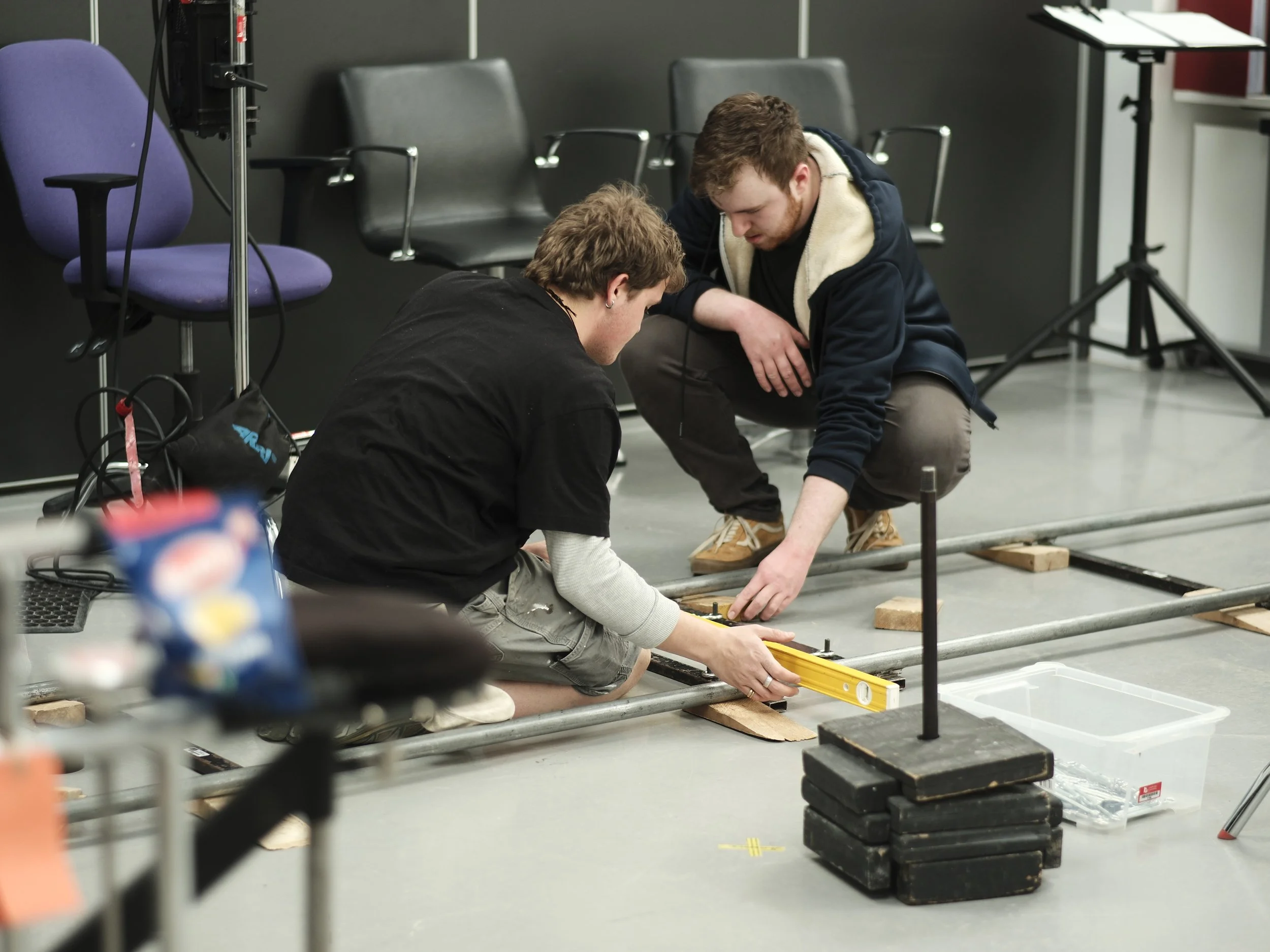 Two young men are working together on a project involving metal pipes on the floor in a room with chairs, a table, and equipment. They are measuring or aligning the pipes, with one using a level and the other adjusting the pipes.