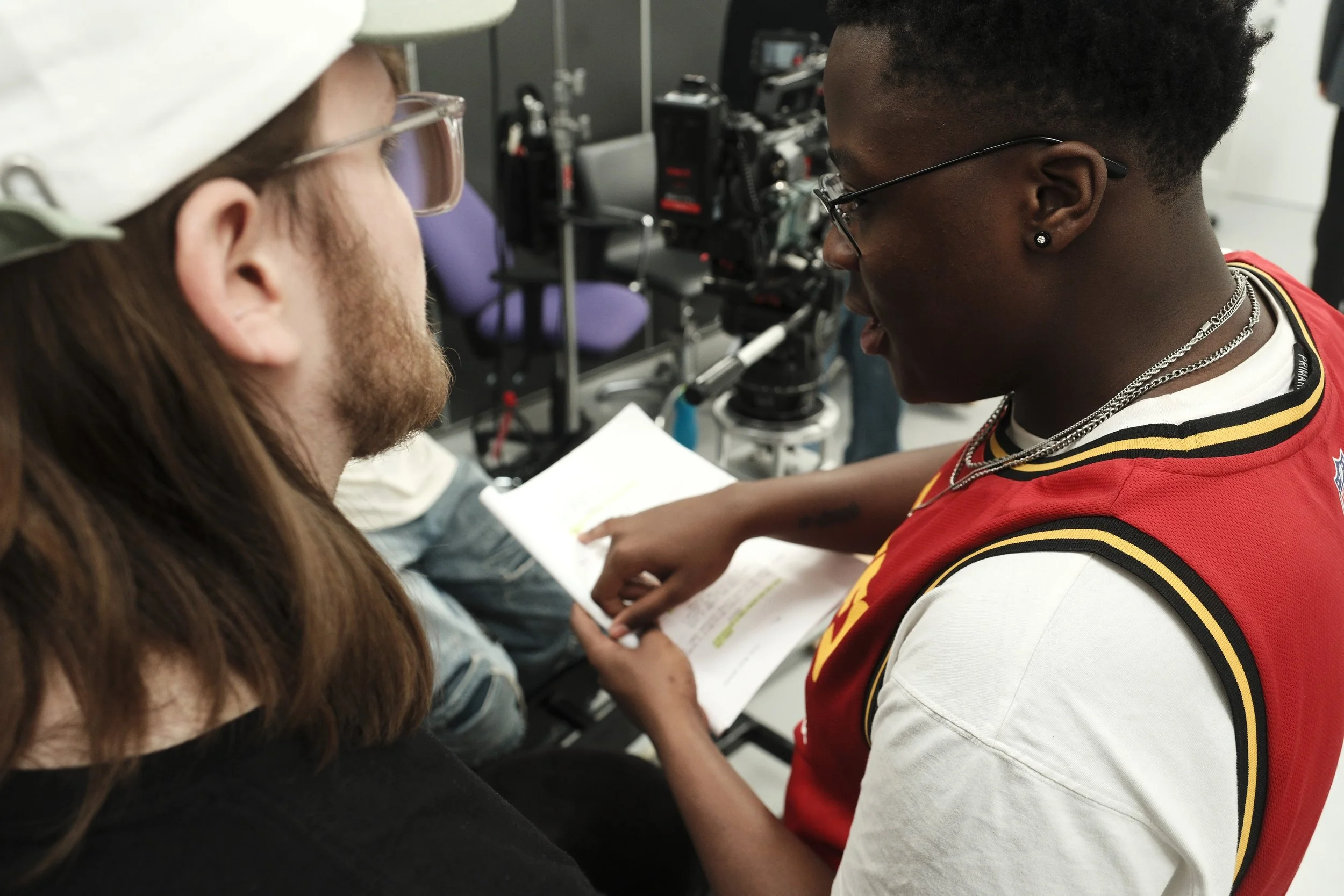 Two young people reviewing a script or notes together, with camera equipment visible in the background.