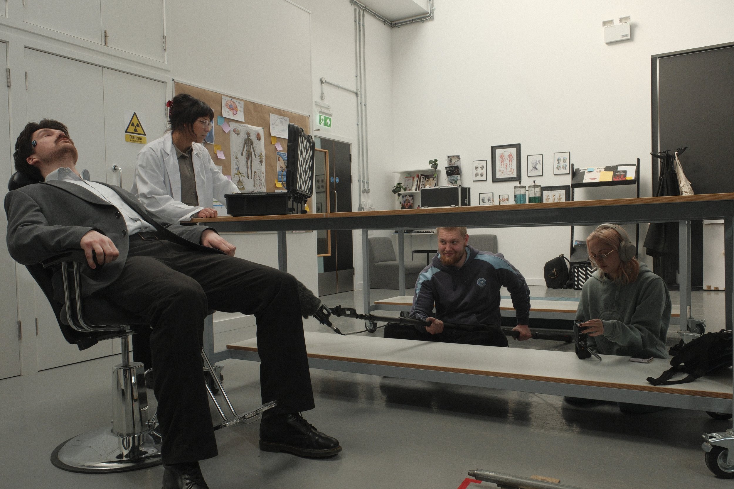 A person is lying in a medical chair with their eyes closed, connected to a chain attached to a person kneeling on the floor, holding the chain. Another person is in the background operating equipment, and a woman with headphones is sitting at the ta