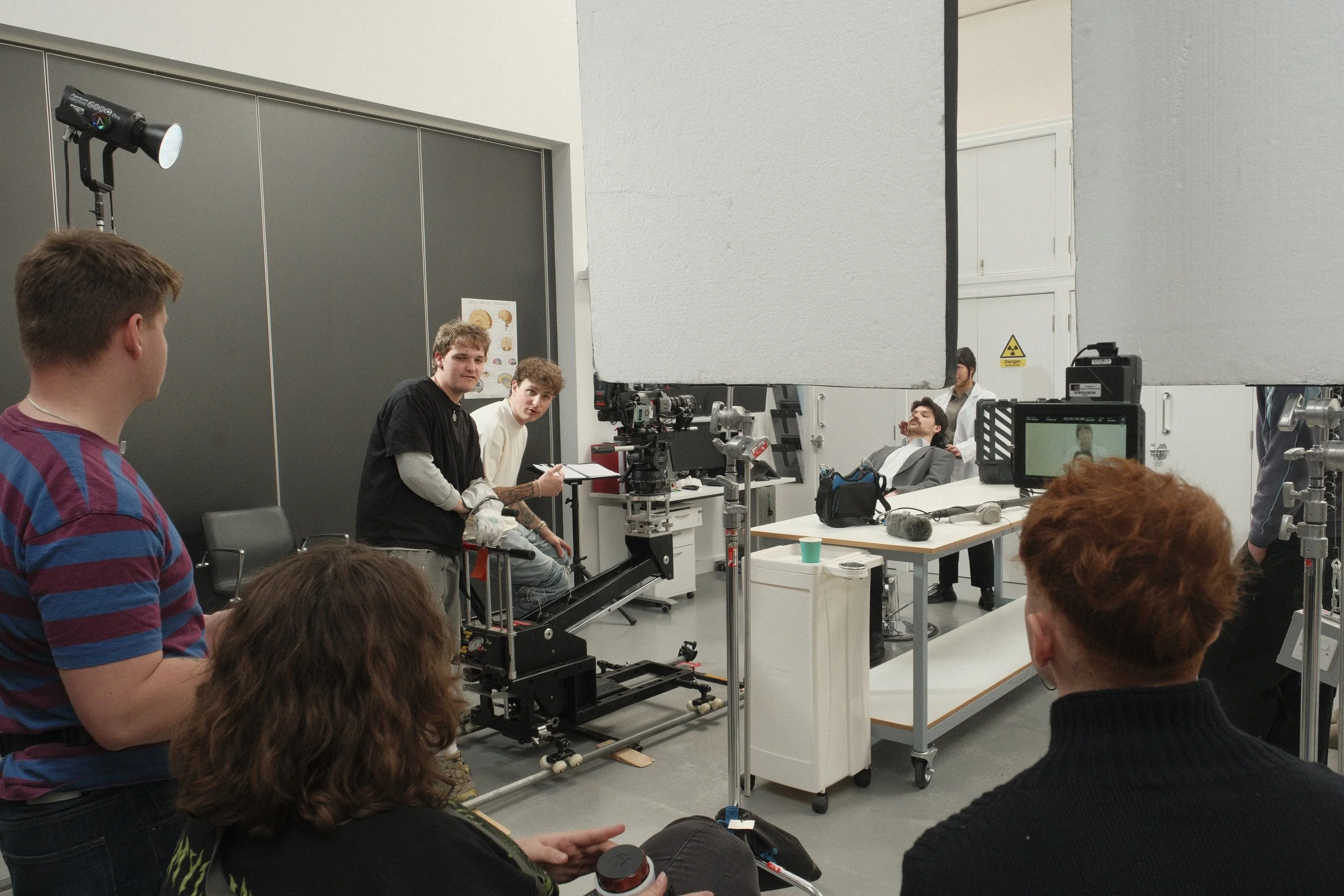 Film crew and actors on a set, with professional camera equipment, large diffusers, and crew members preparing for a scene involving a man sitting at a desk.