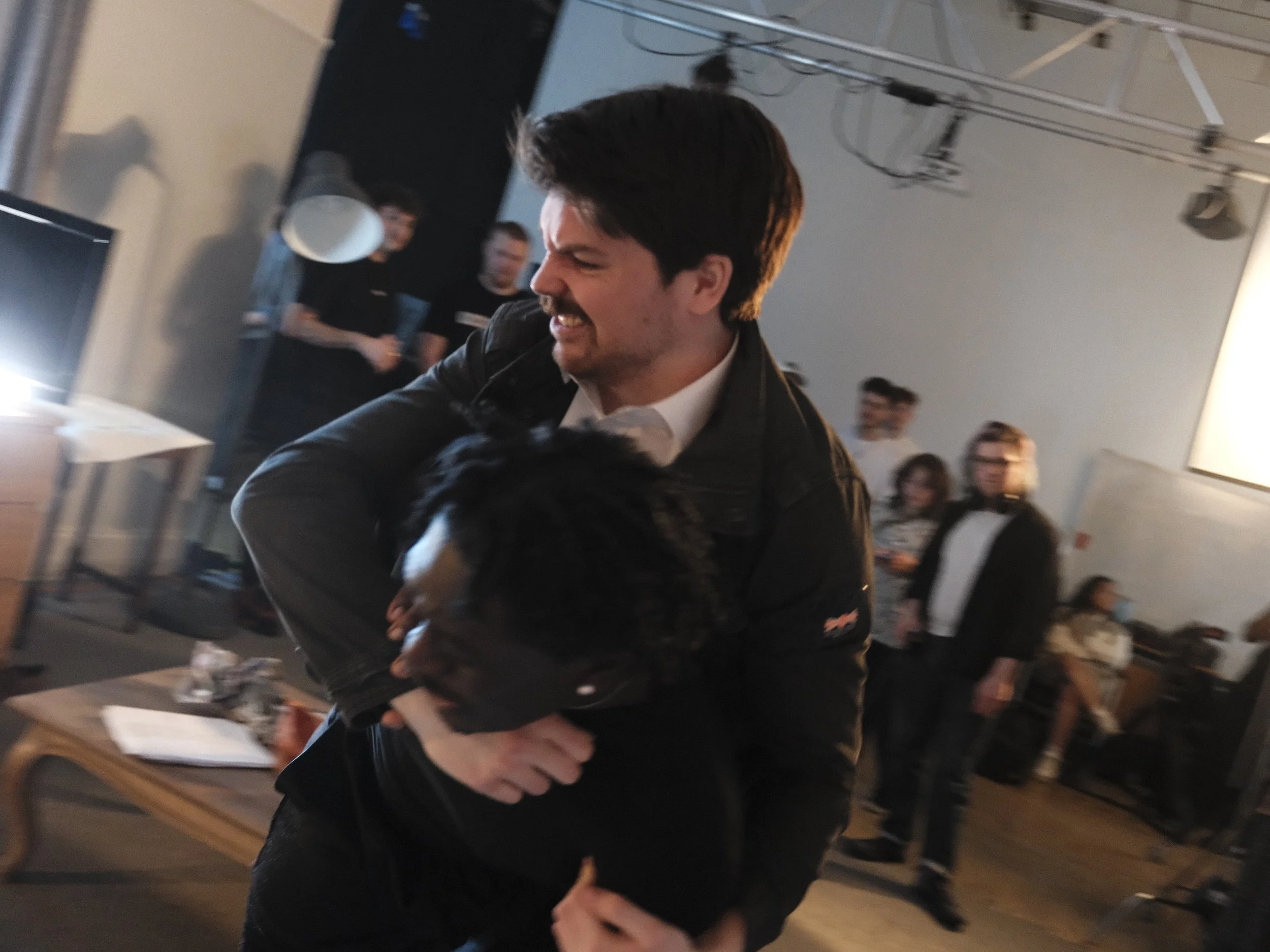 A man with dark hair and a beard, wearing a black jacket and white shirt, smiling and hugging a black and white dog in an indoor setting with several people in the background.