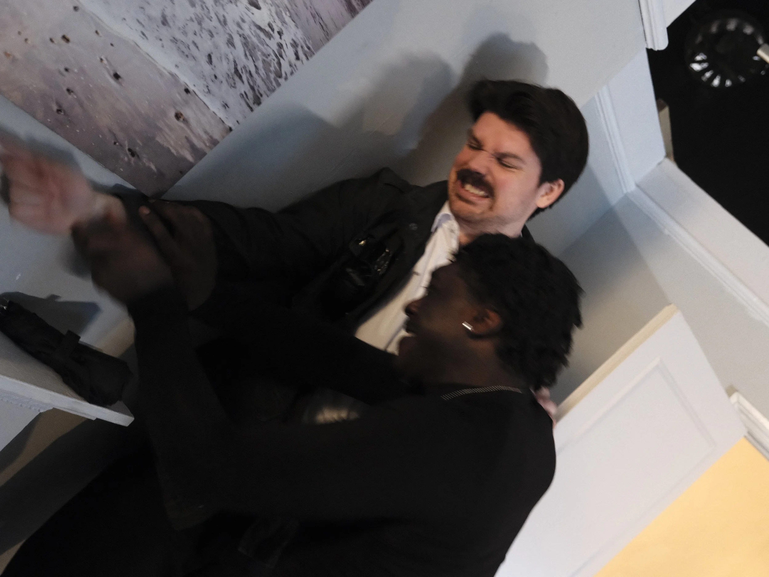 A man is sitting on the floor, grimacing in pain or distress, while a woman stands over him, holding his hand or arm. The setting appears to be indoors, near a wall and a door.