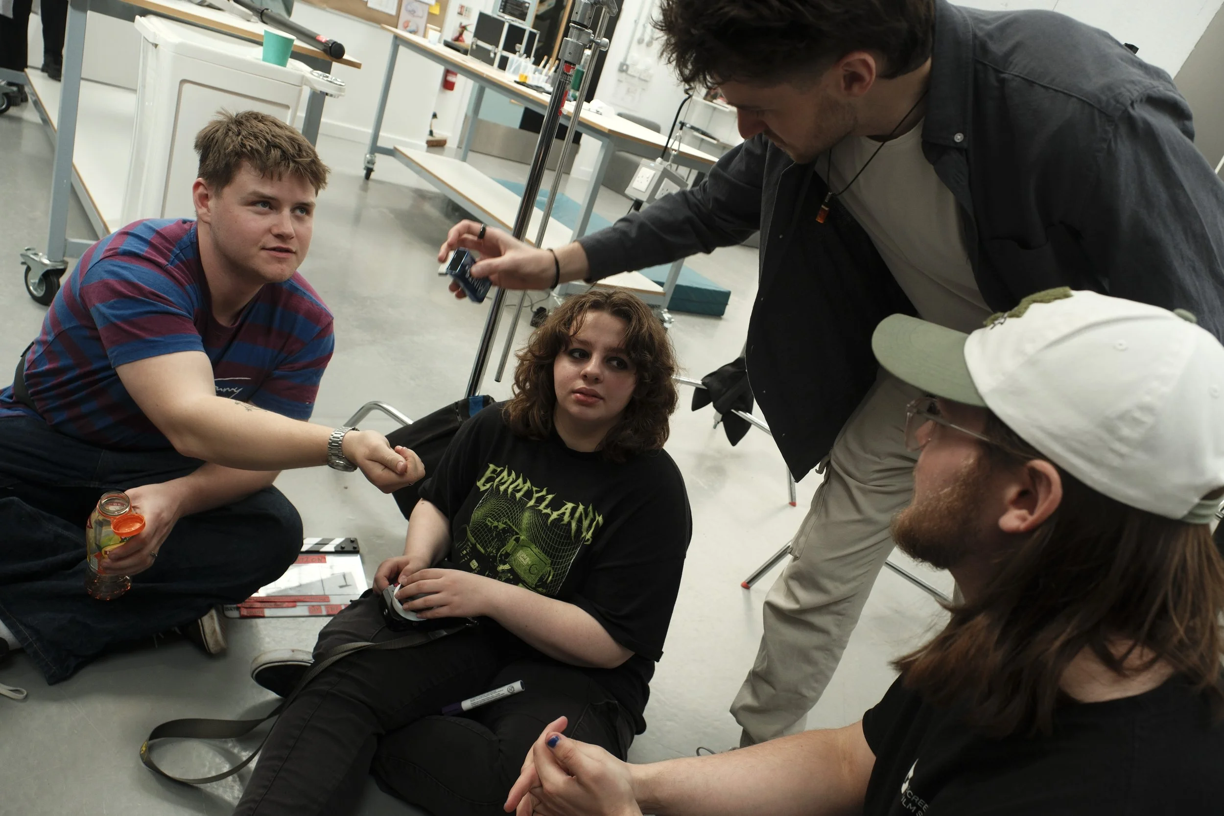 Four young people having a discussion in a classroom or workshop setting, with one person sitting on the floor and others standing or kneeling around him, engaging in a conversation.