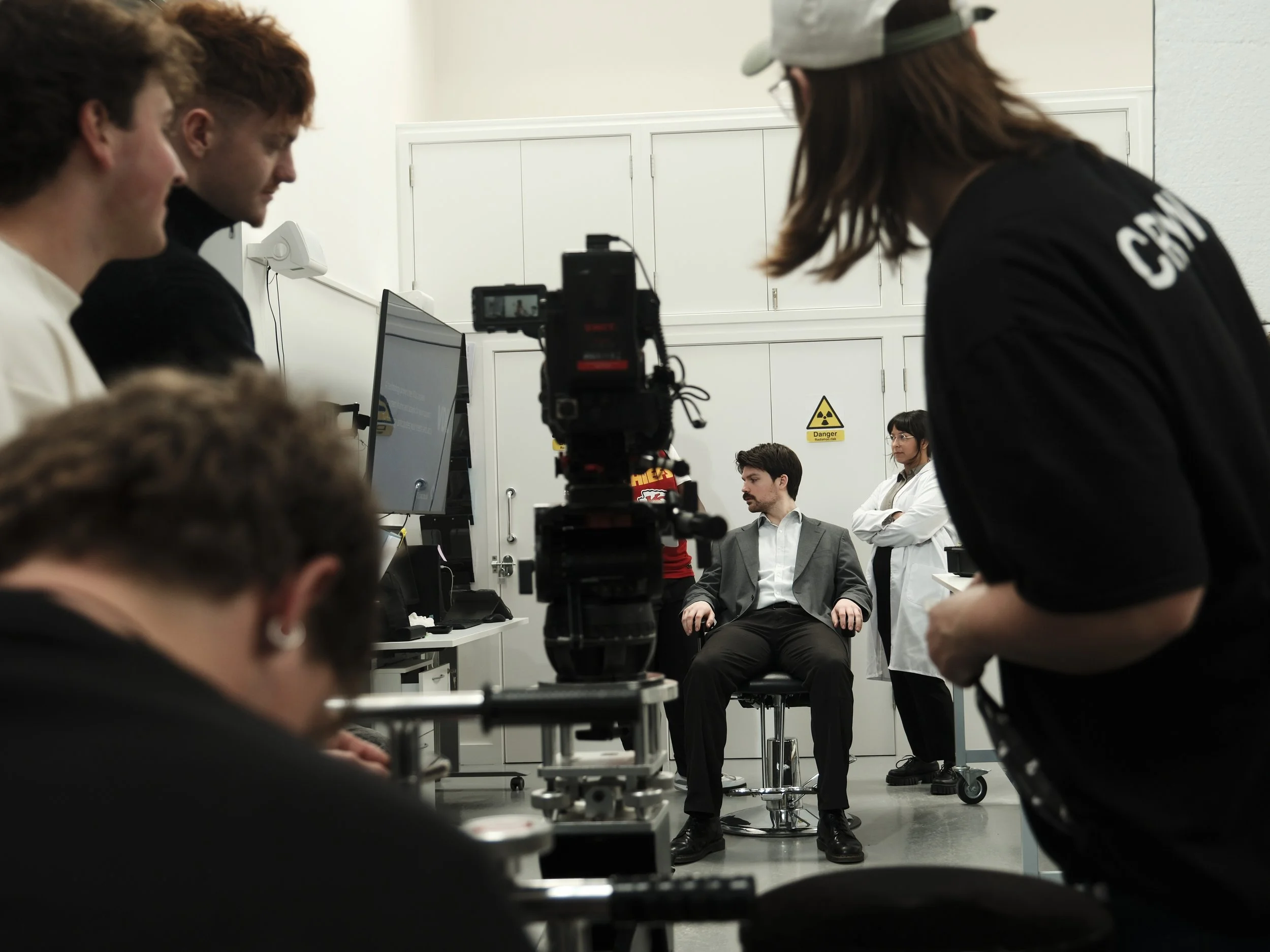 A film crew is filming a scene in a laboratory with a man in a suit sitting on a chair, a woman in a lab coat standing with arms crossed, and another person operating equipment. A warning sign with a radiation symbol is visible on the door in the bac