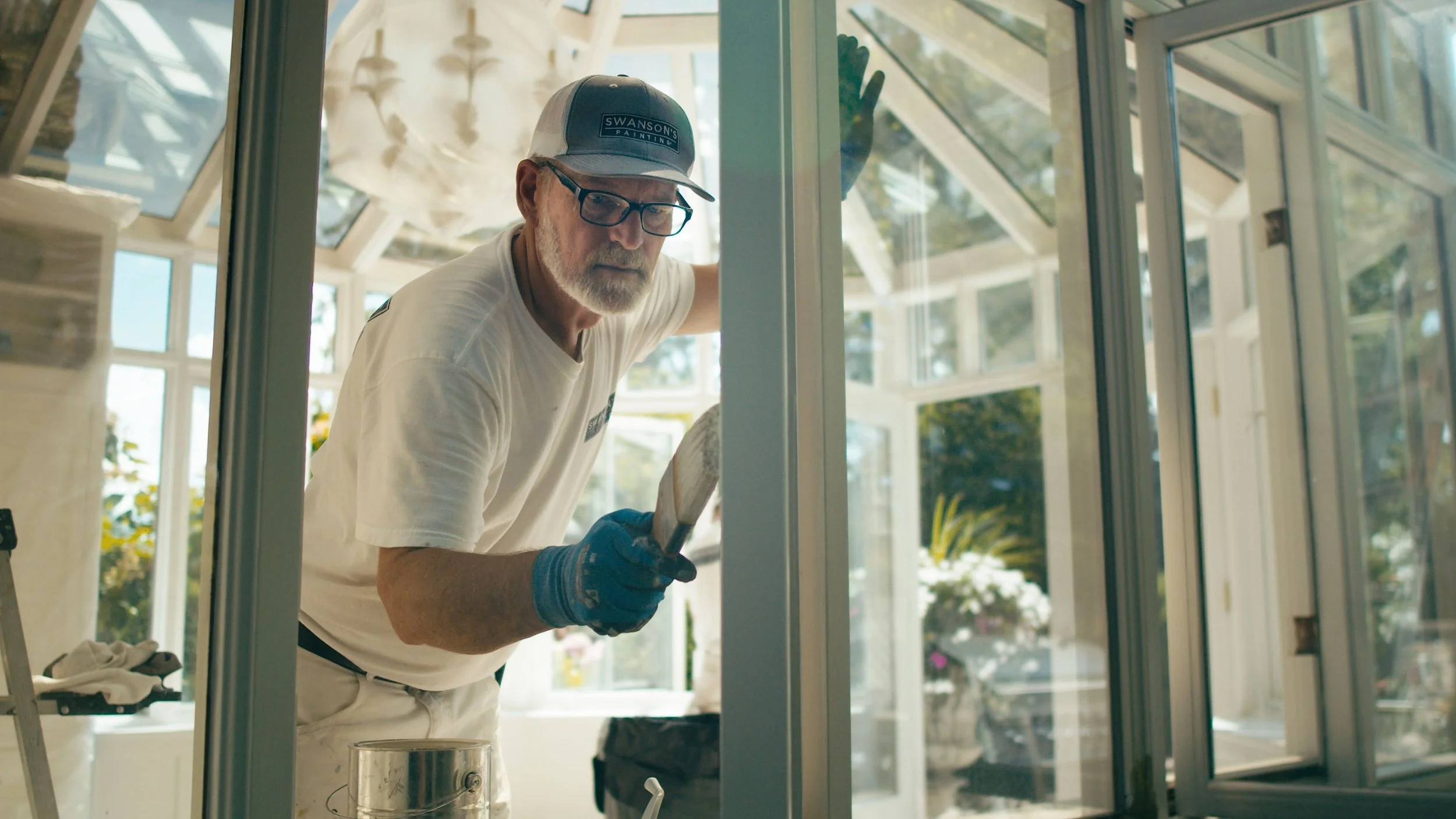 An elderly man wearing glasses, a cap, and blue gloves is painting on a window frame inside a bright, sunny room.