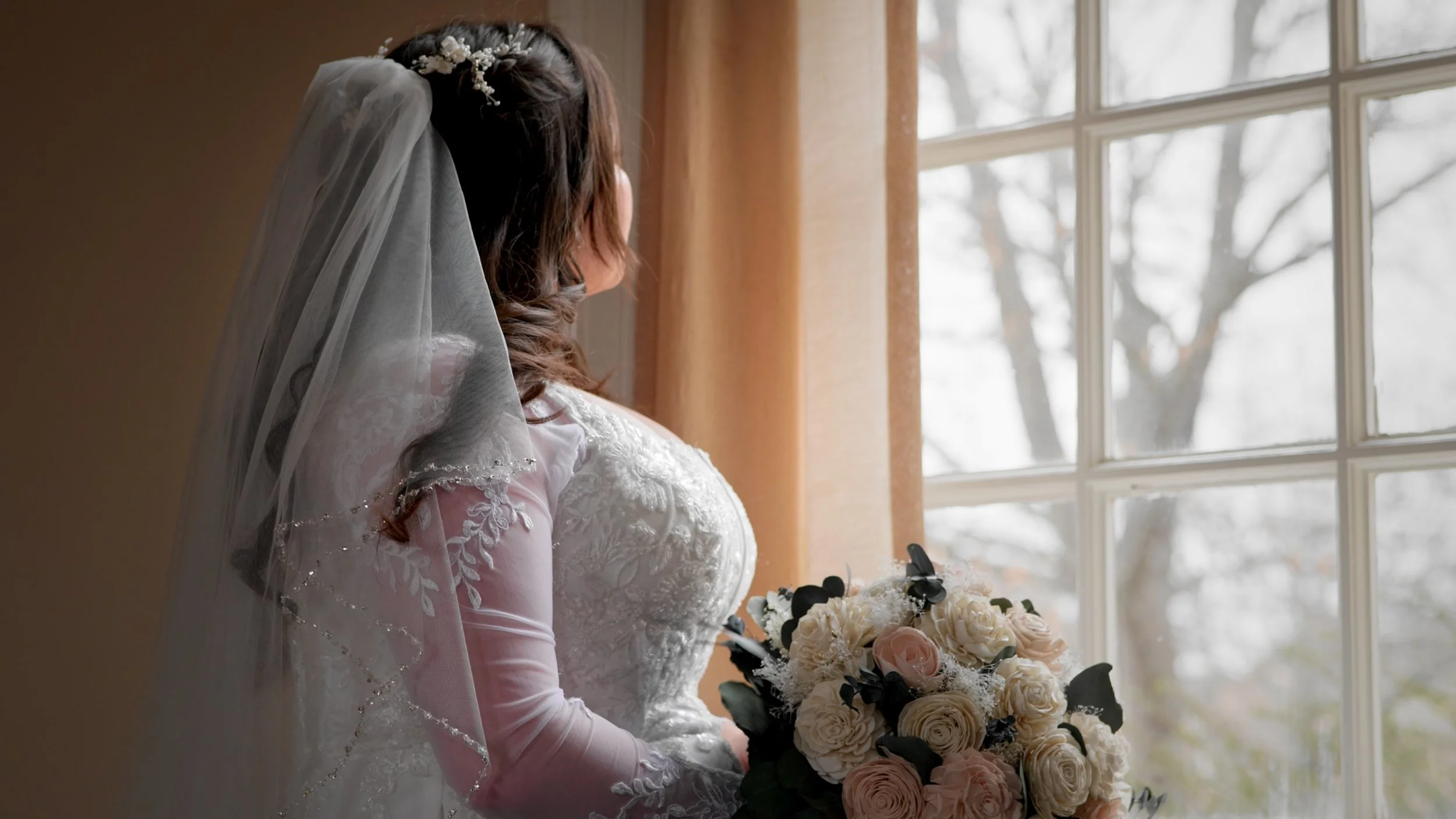 Brunette bride in wedding dress with veil looking out a window, holding a bouquet of white and pink roses.