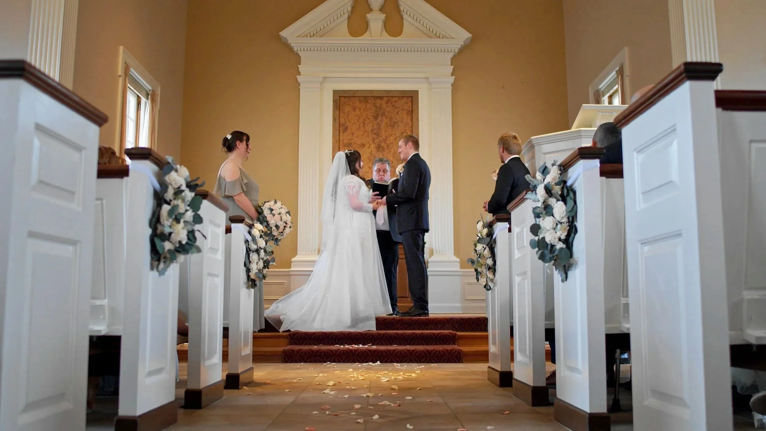 A wedding ceremony in a church with a bride and groom holding hands and exchanging vows, officiant in the background, bridesmaid and groomsman standing beside them, decorated pews with flowers and ribbons, and flower petals on the aisle.